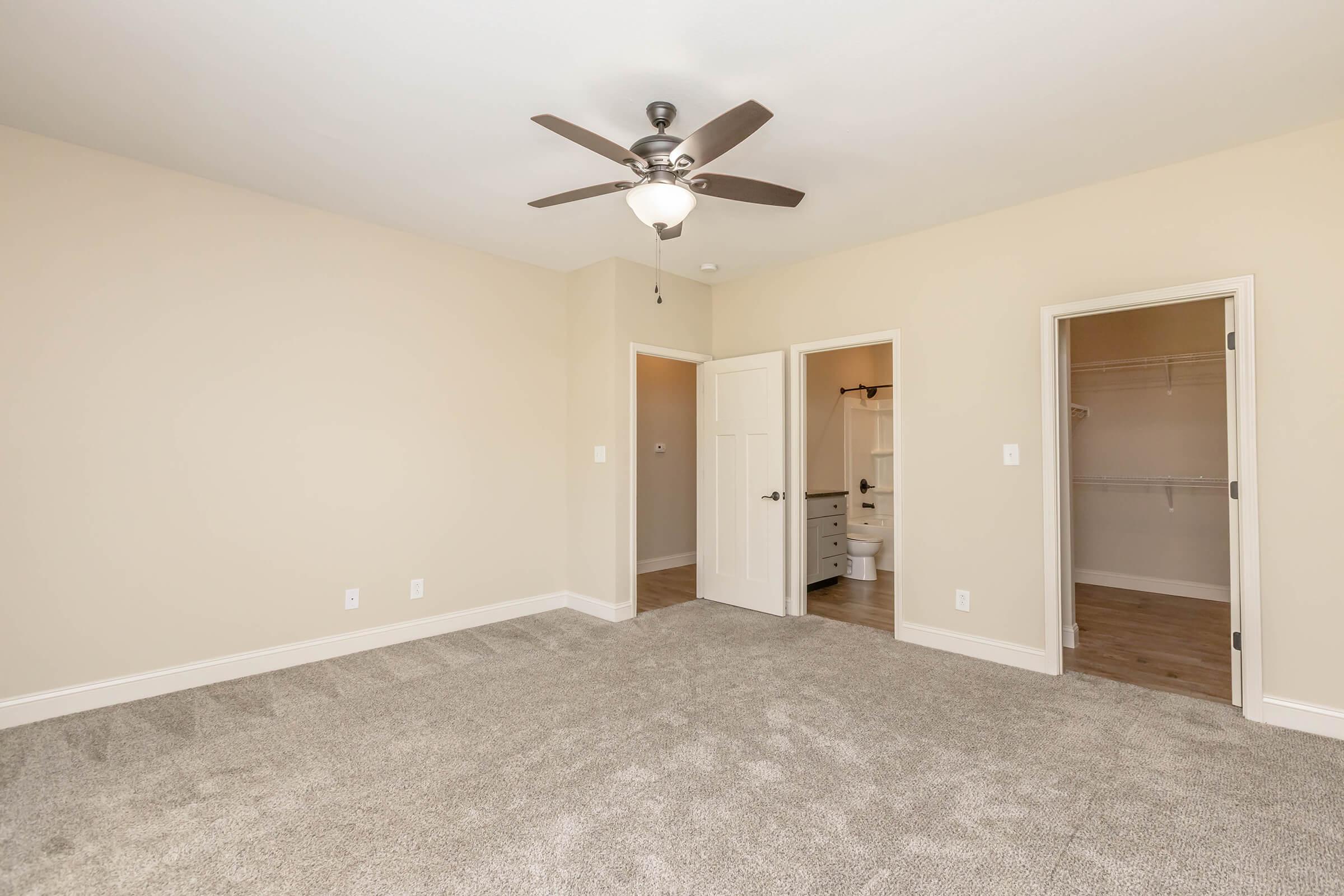 A spacious, empty bedroom with light beige walls and a ceiling fan. The floor is covered with carpet, and there are two doorways leading to a bathroom and a closet. Natural light from a window brightens the room, creating a clean and inviting atmosphere.