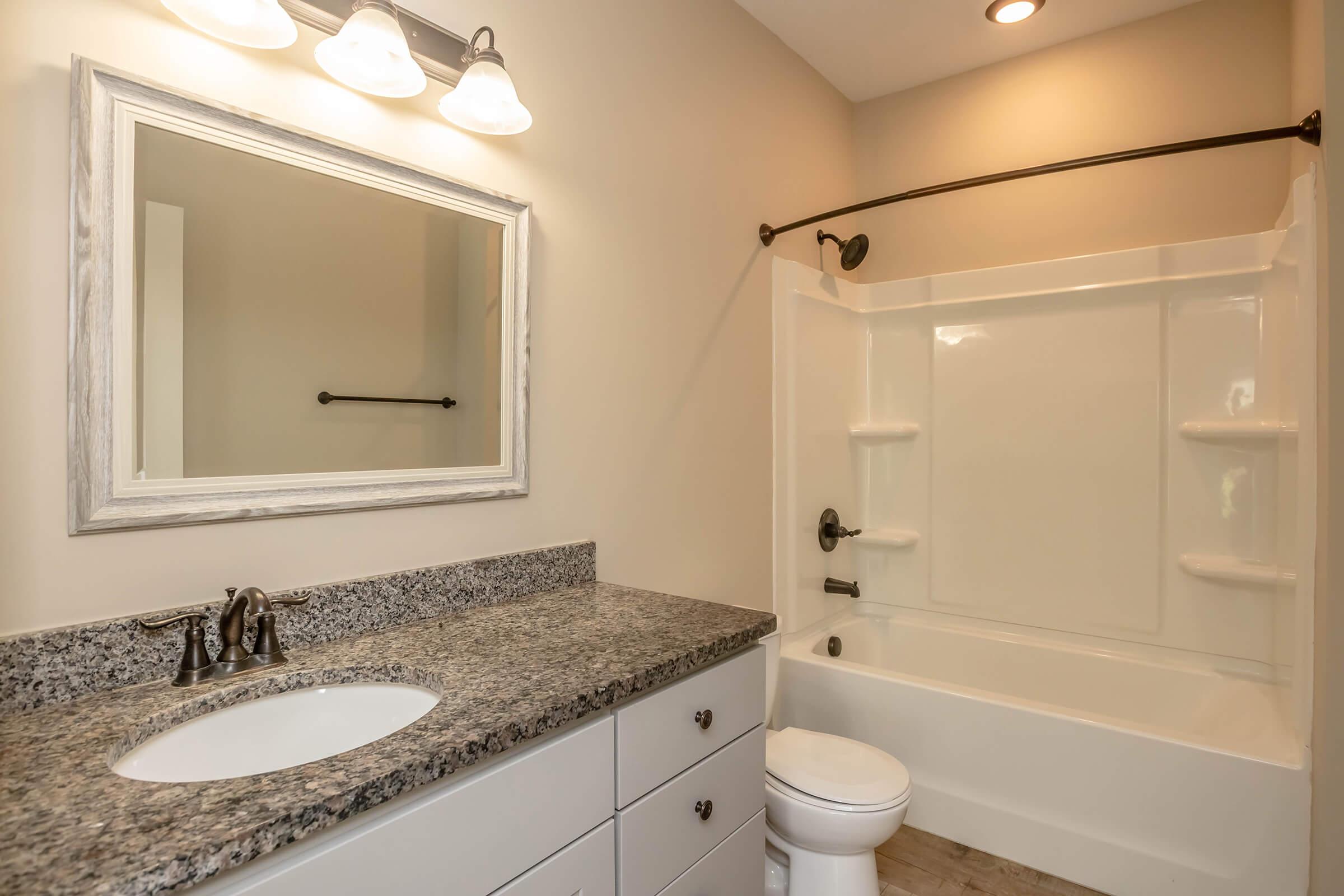 A modern bathroom featuring a white bathtub with a shower, a granite countertop with a sink, and a large mirror framed in white. The walls are painted in a neutral tone, and there are three light fixtures above the mirror. The flooring consists of light-colored tiles.
