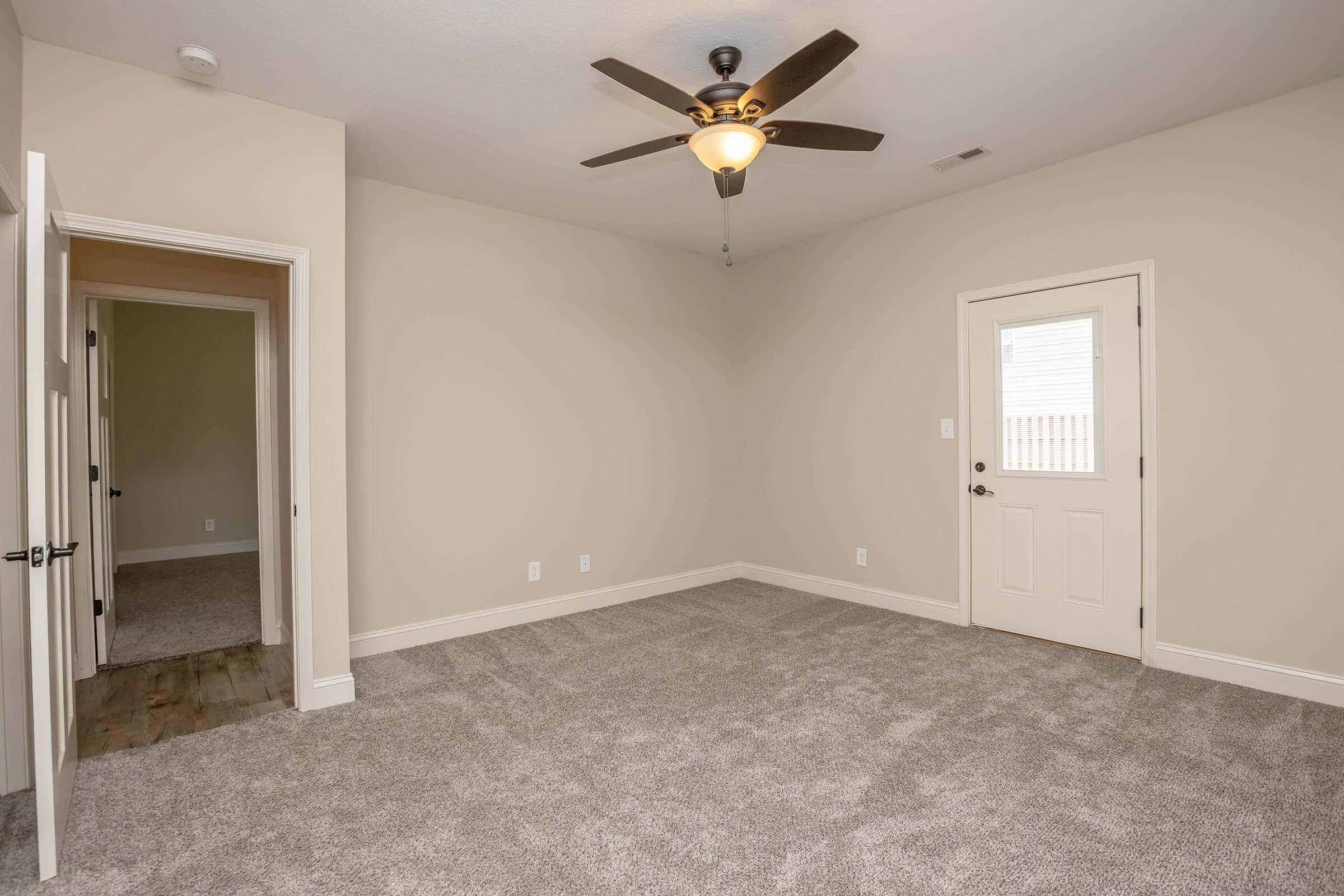 A spacious, empty room with light-colored walls and plush carpet. It features a ceiling fan with a light fixture. A door leads outside, while another door leads to a hallway. The room is well-lit with natural light, creating an inviting atmosphere.