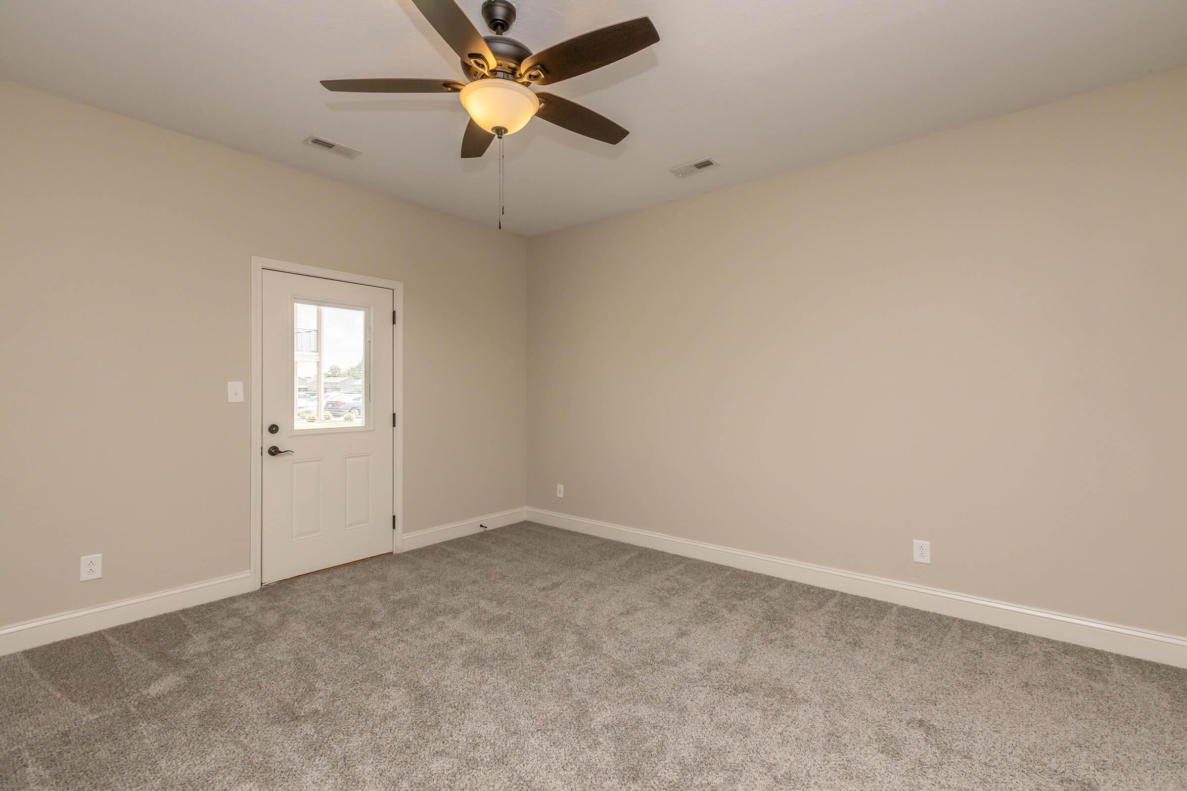 A spacious, empty room with light beige walls and plush gray carpet. A ceiling fan with wooden blades is installed overhead. There is a closed door with a glass panel on the left side, providing access to the exterior. Natural light enters through the door, giving the room a bright atmosphere.