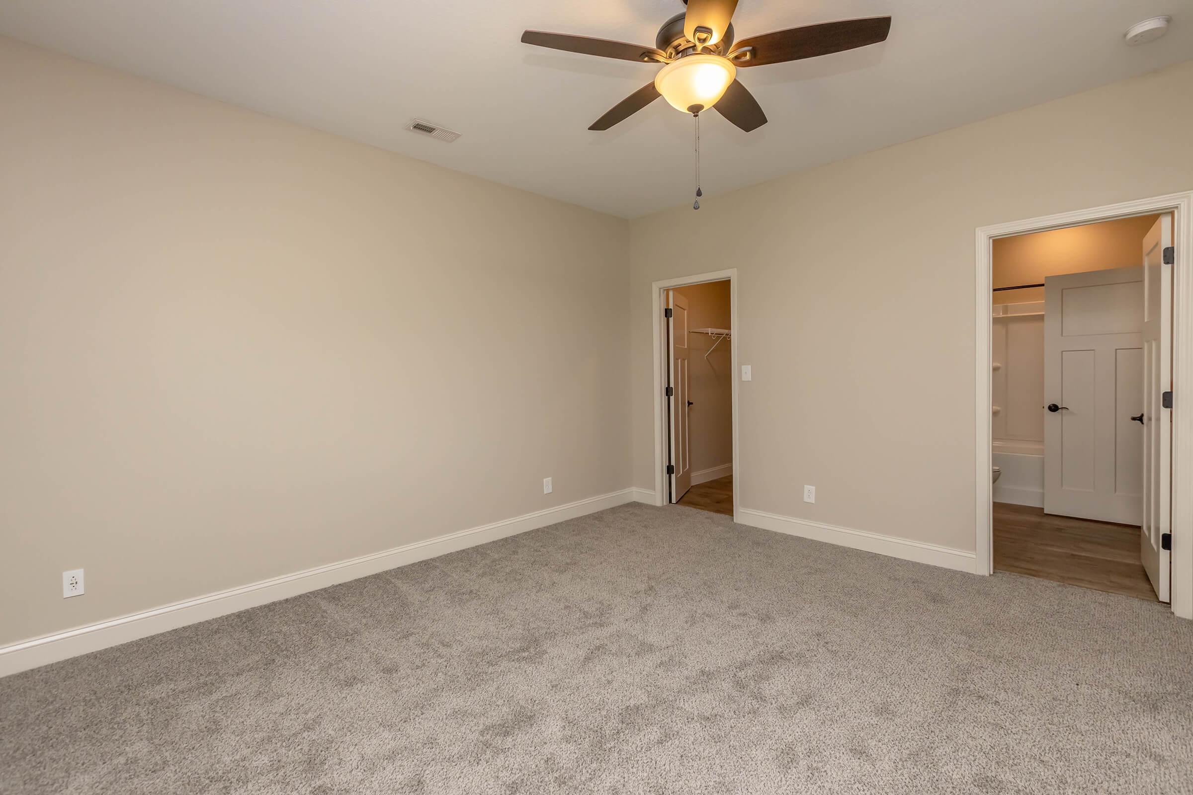 Empty room with light beige walls and plush gray carpet. A ceiling fan with light is mounted on the ceiling, and there are two doorways: one leading to a closet and the other to a bathroom. The space is well-lit and features neutral tones, creating a clean and inviting atmosphere.
