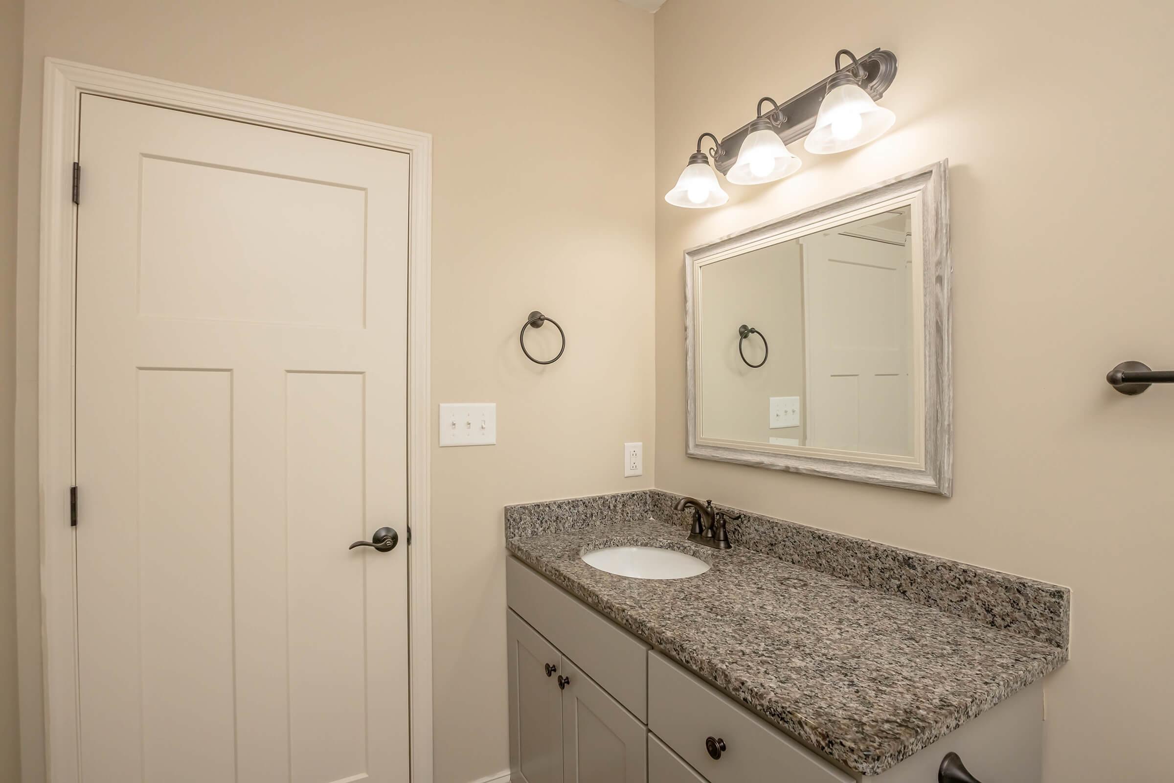 A modern bathroom featuring a light-colored wall, a granite countertop with a dark faucet, a mirror framed in wood, and three light fixtures above it. The room also has a white door and a towel holder mounted on the wall.