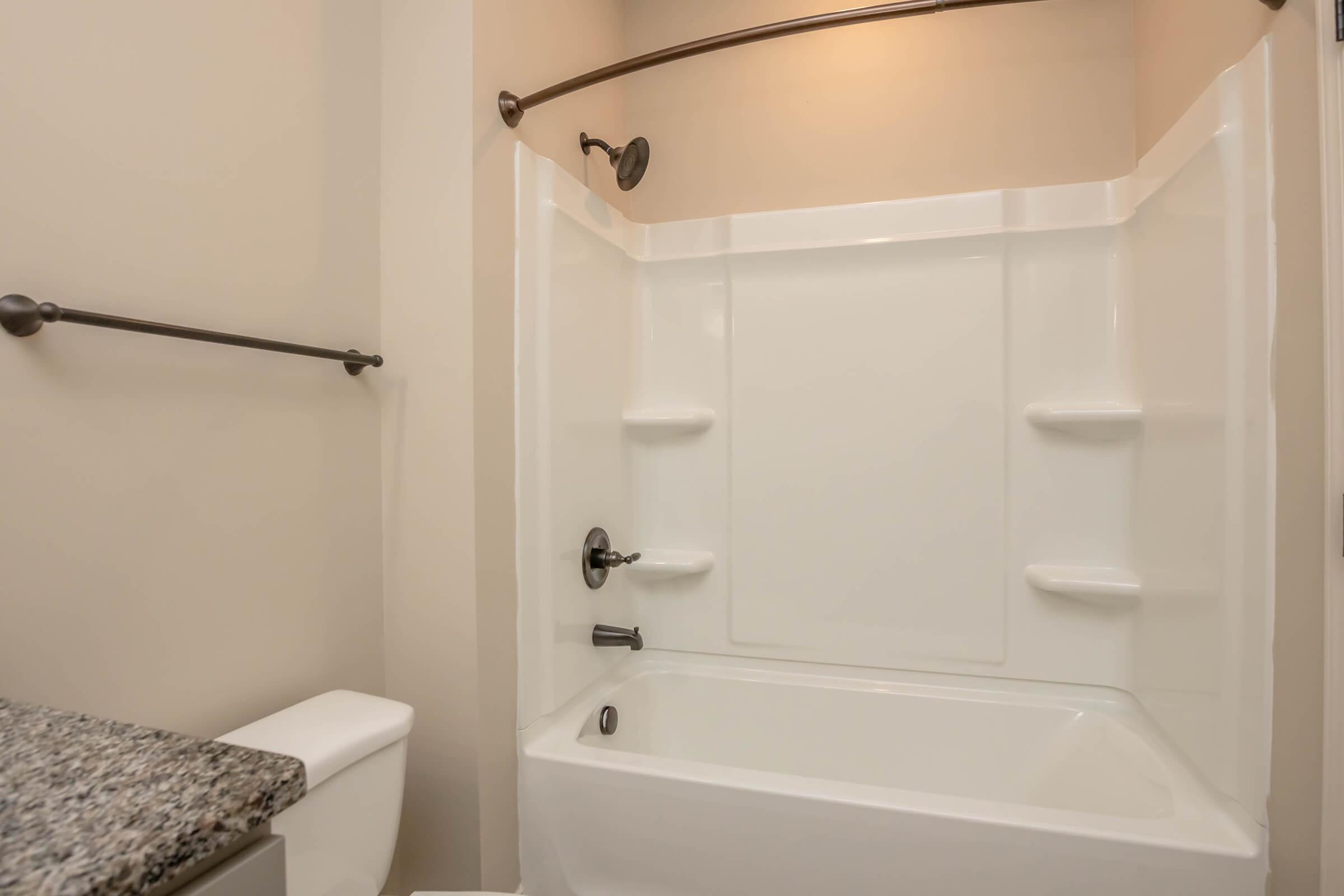 A clean, modern bathroom featuring a white bathtub and shower combination with built-in shelves. The walls are a neutral color, and there's a granite countertop visible beside a white toilet. The space is well-lit with a light fixture above the shower area.