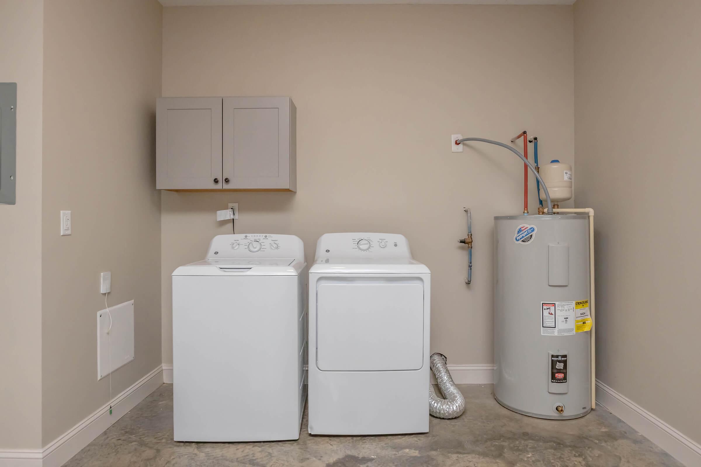 A clean laundry room containing a white washing machine and dryer side by side, with light gray cabinets above. To the right, there is a gray water heater with a connected vent. The walls are a light beige color, and the flooring appears to be a textured concrete.
