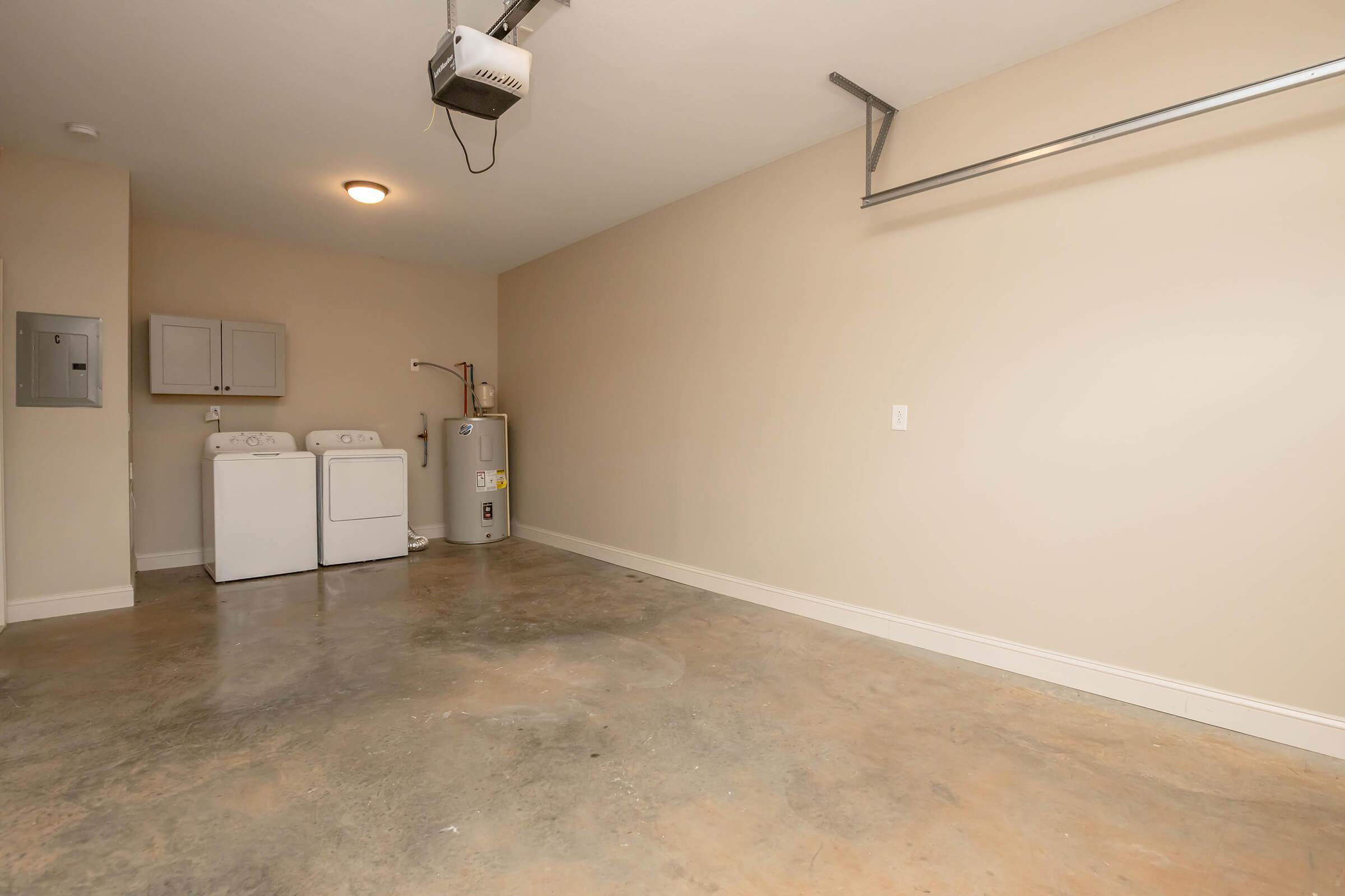 A spacious garage featuring a washer and dryer set against a neutral-colored wall. The floor is polished concrete, and there is a ceiling-mounted projector. A utility area is visible on the left with a service panel. The overall space is clean and well-lit.