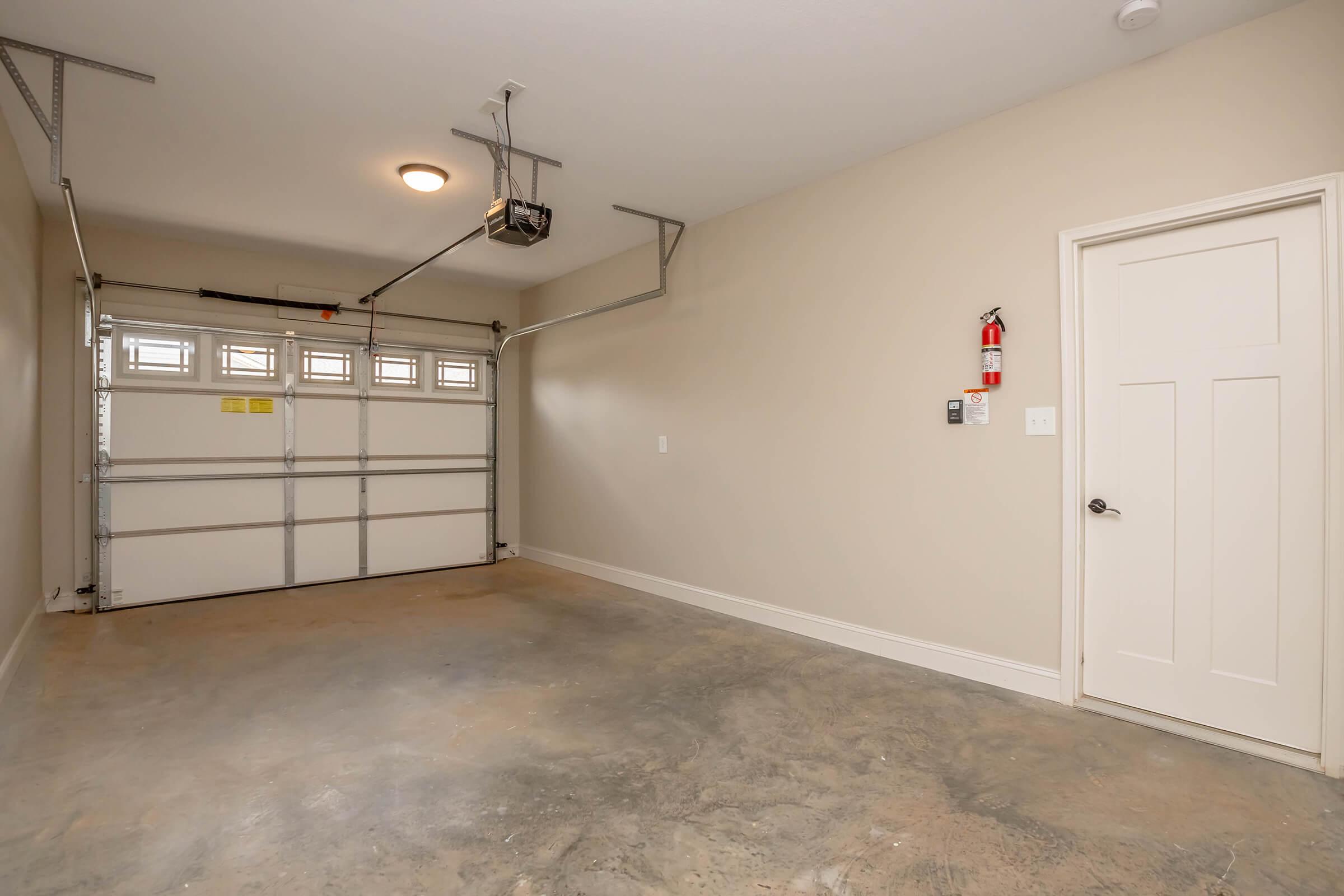 A spacious, empty garage with a gray concrete floor, featuring a white garage door with windows, an overhead light fixture, and a fire extinguisher mounted on the wall near a closed door. The walls are painted in a light beige color.
