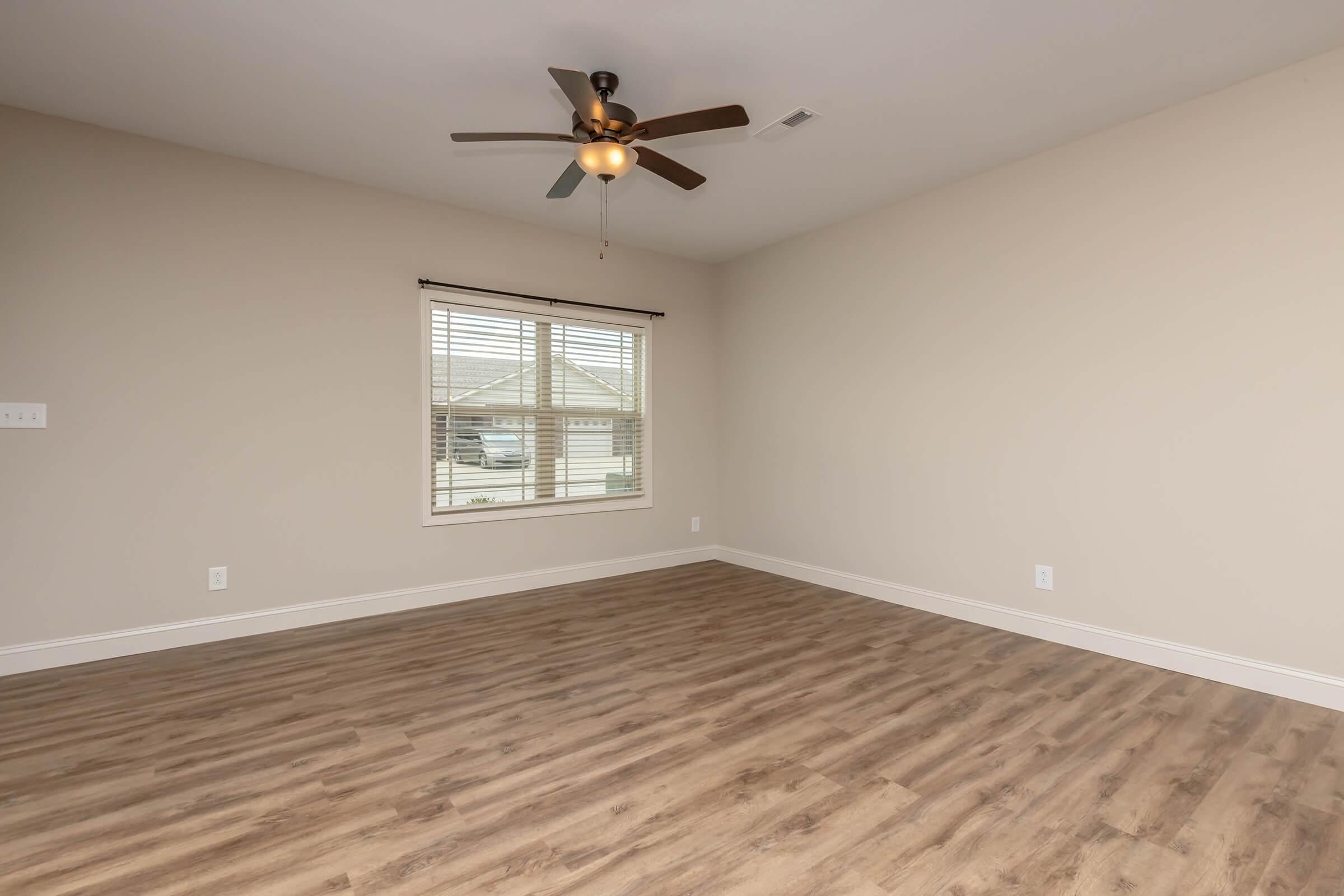 A spacious, empty room with light beige walls and a ceiling fan. It features a large window with blinds that allows natural light to enter. The floor is made of light-colored laminate wood. The room has a clean and modern aesthetic, suitable for various uses.