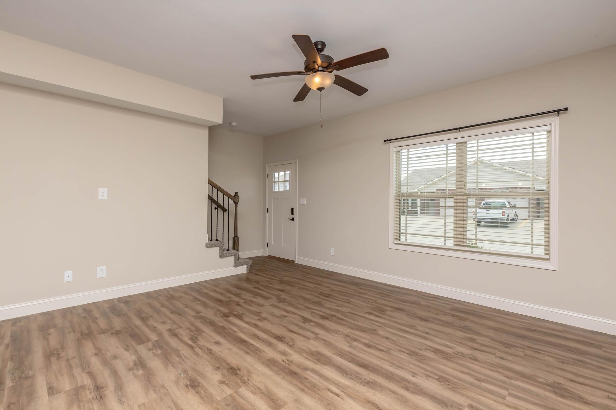 Spacious living room featuring light-colored walls, a ceiling fan, and a large window with blinds. The room has wood-style flooring and a staircase leading to the upper level. A front door is visible, providing access to the outside. Natural light brightens the area, creating an inviting atmosphere.