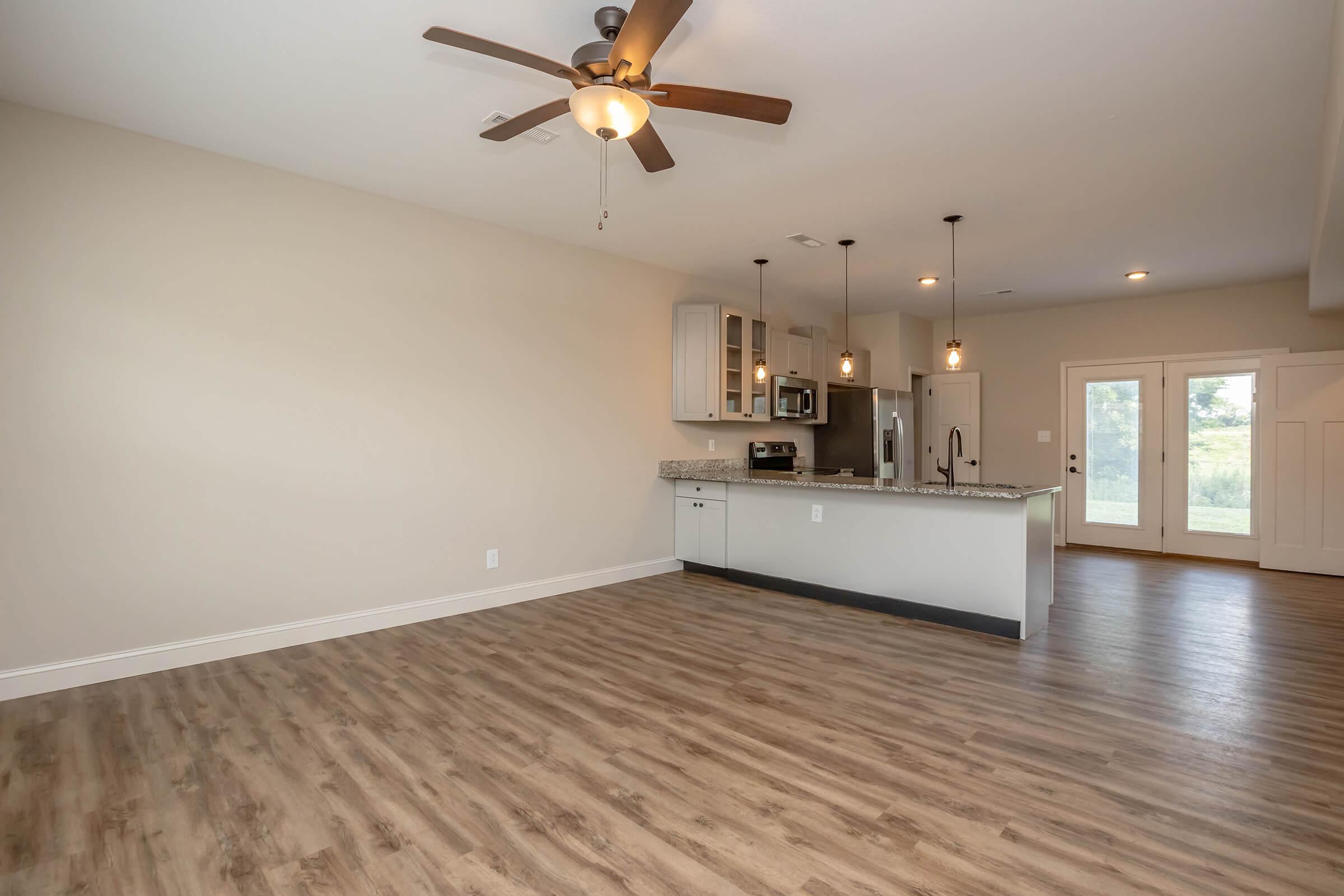 A spacious modern living area featuring light wood-look flooring, a ceiling fan, and an open kitchen. The kitchen includes a granite countertop, stainless steel appliances, and pendant lighting. Large windows and a door provide access to an outdoor space, creating a bright and airy atmosphere.