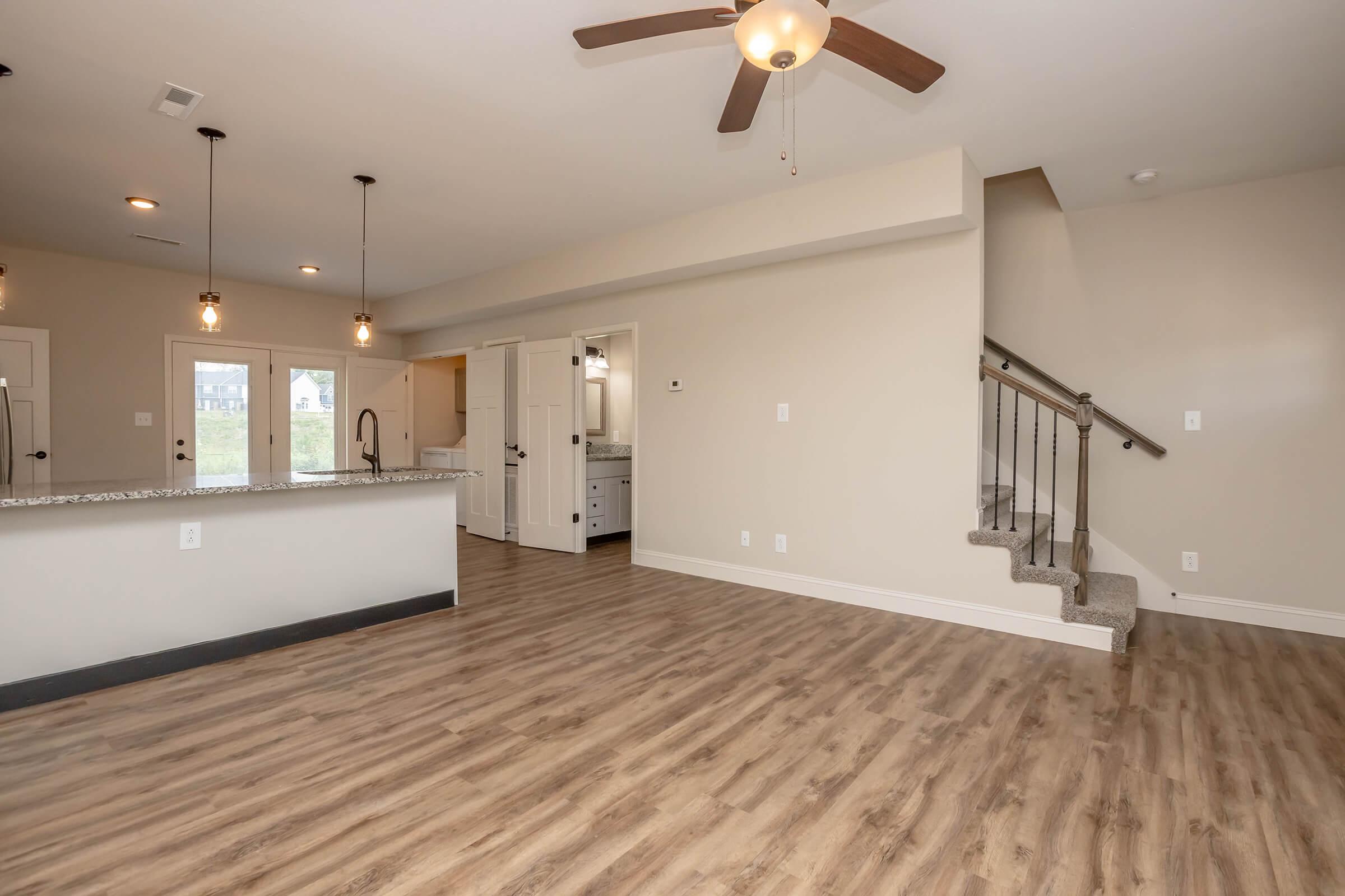 Interior of a modern home featuring an open floor plan. The living area has wooden flooring and a ceiling fan. There is a kitchen island with pendant lighting and a staircase leading to the upper level. Soft beige walls and a doorway leading to additional rooms are visible.