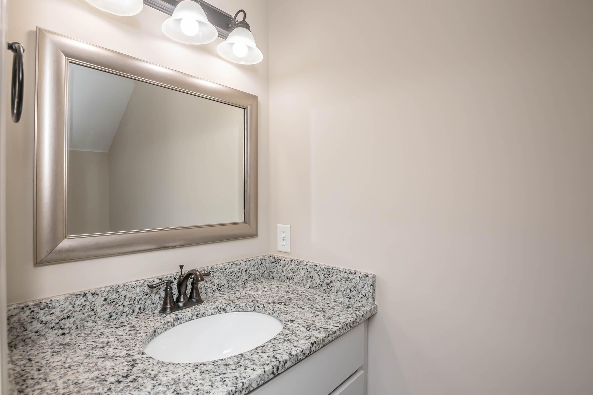 A clean bathroom with a granite countertop, featuring a sink and faucet. Above the sink, there is a large, framed mirror with three light fixtures. The wall is painted in a neutral color, enhancing the modern and simplistic design of the space.