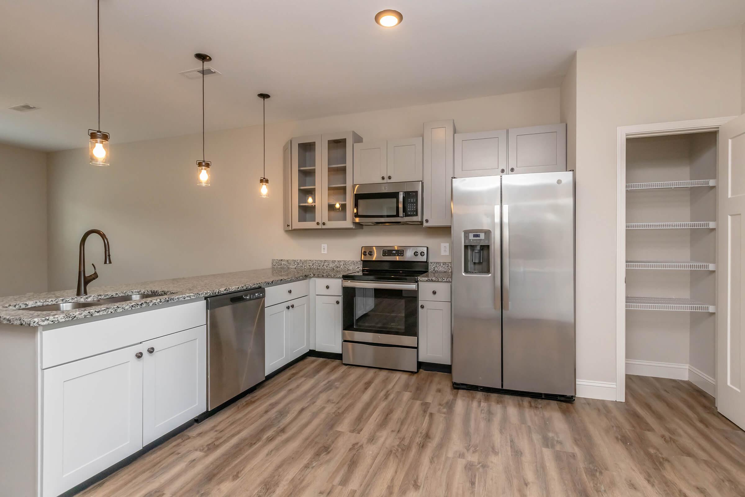 Modern kitchen featuring stainless steel appliances including a refrigerator, microwave, and oven. The granite countertops complement white cabinets, and there are hanging light fixtures above. The floor is made of light-colored wood, and there is a pantry adjacent to the kitchen with an open shelving design.