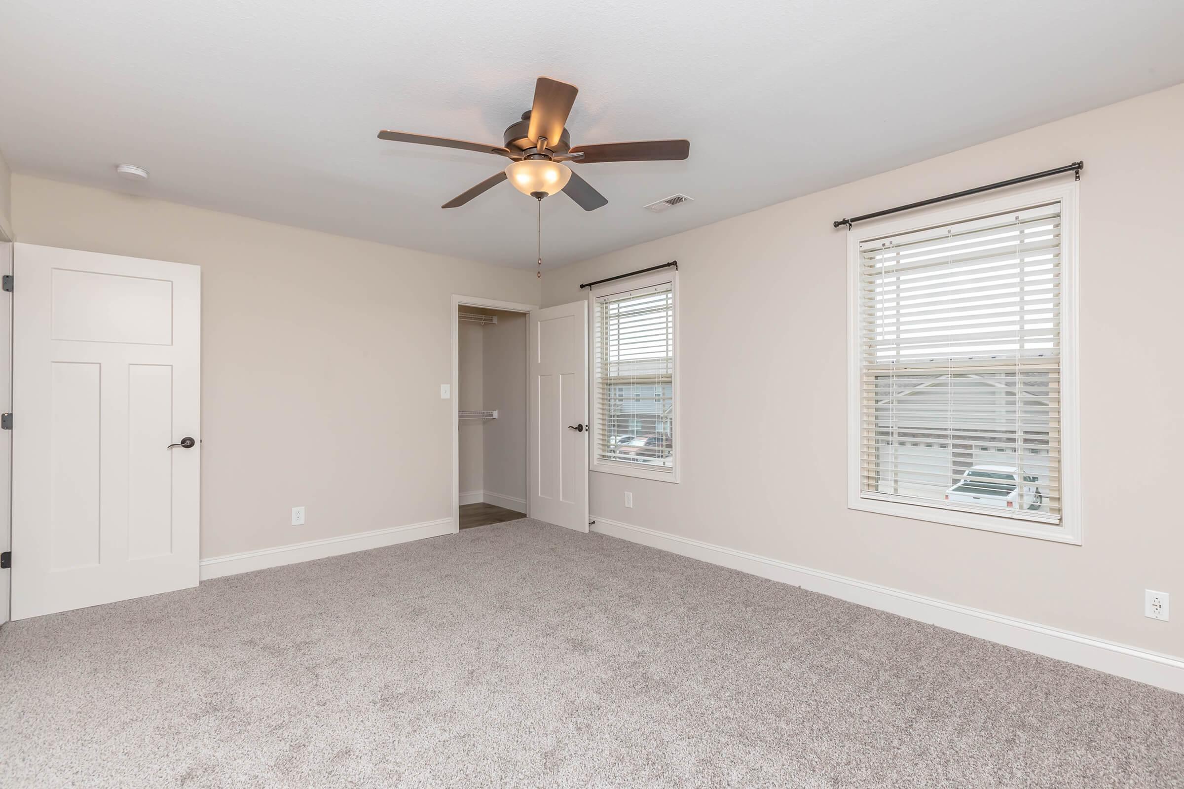 A well-lit, empty bedroom featuring light beige walls, new gray carpeting, and a ceiling fan with five blades. Two windows with white blinds allow natural light, and a closed door leads to another room. The overall space is clean and modern, ready for decoration.