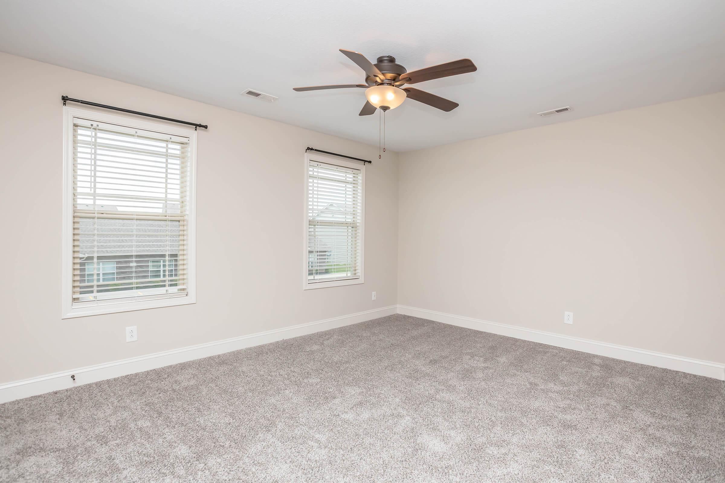 Empty room with light-colored walls and light gray carpet. Two windows with white blinds allow natural light. A ceiling fan with wooden blades is centered in the room. The space is airy and spacious, giving a neutral and inviting atmosphere.