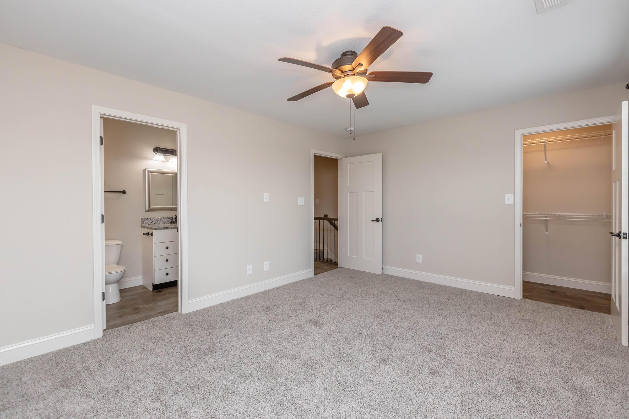 A spacious bedroom with light-colored walls and carpet. It features a ceiling fan, an open closet, and two doors leading to a bathroom and a staircase. Natural light fills the room, highlighting its modern design.