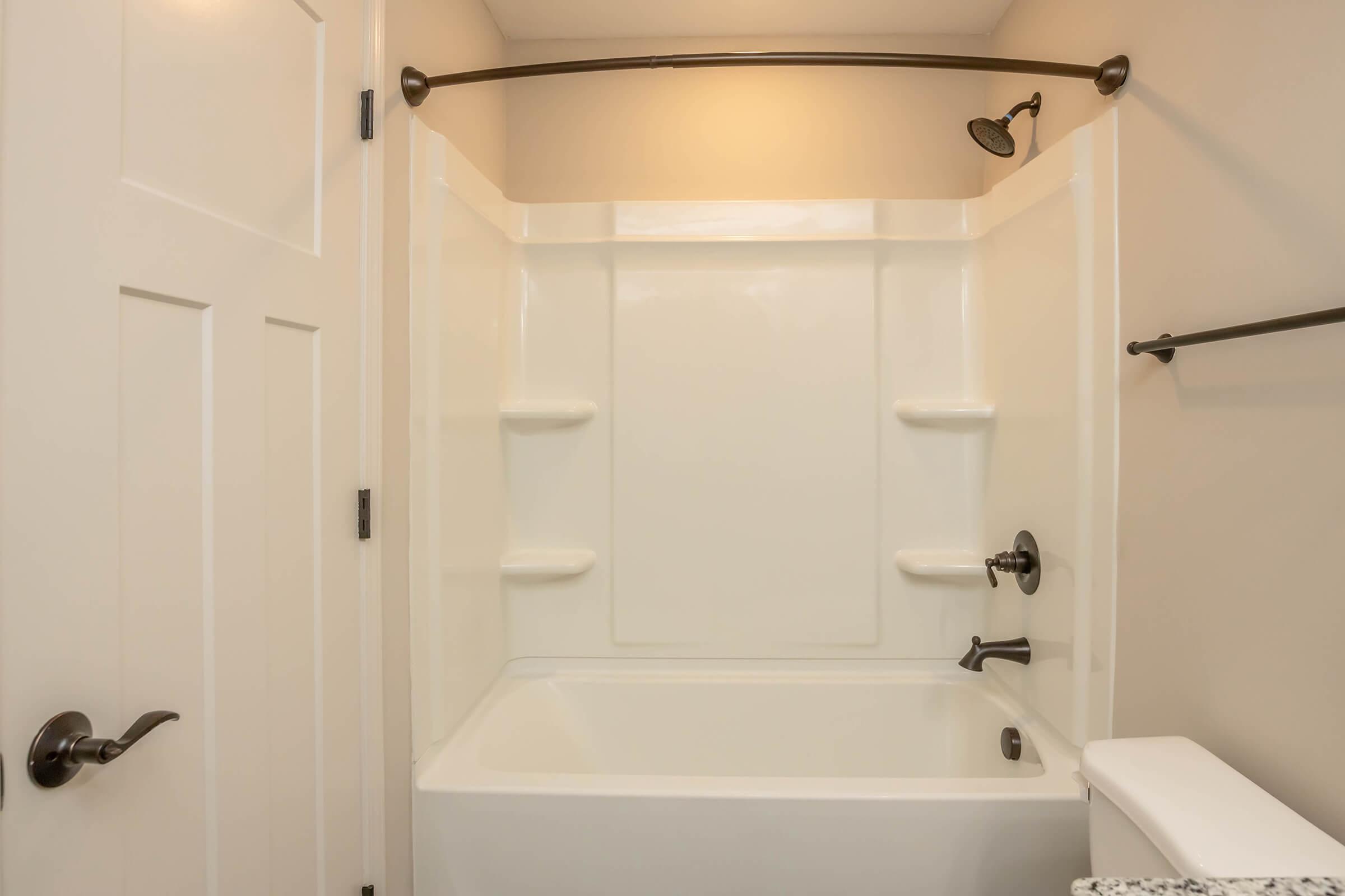 A clean, modern bathroom with a bathtub and shower. The showerhead is mounted on the wall above the tub, and there are two built-in shelves for toiletries. The walls are a light color, and the floor is partially visible, along with a white toilet and a door in the background.