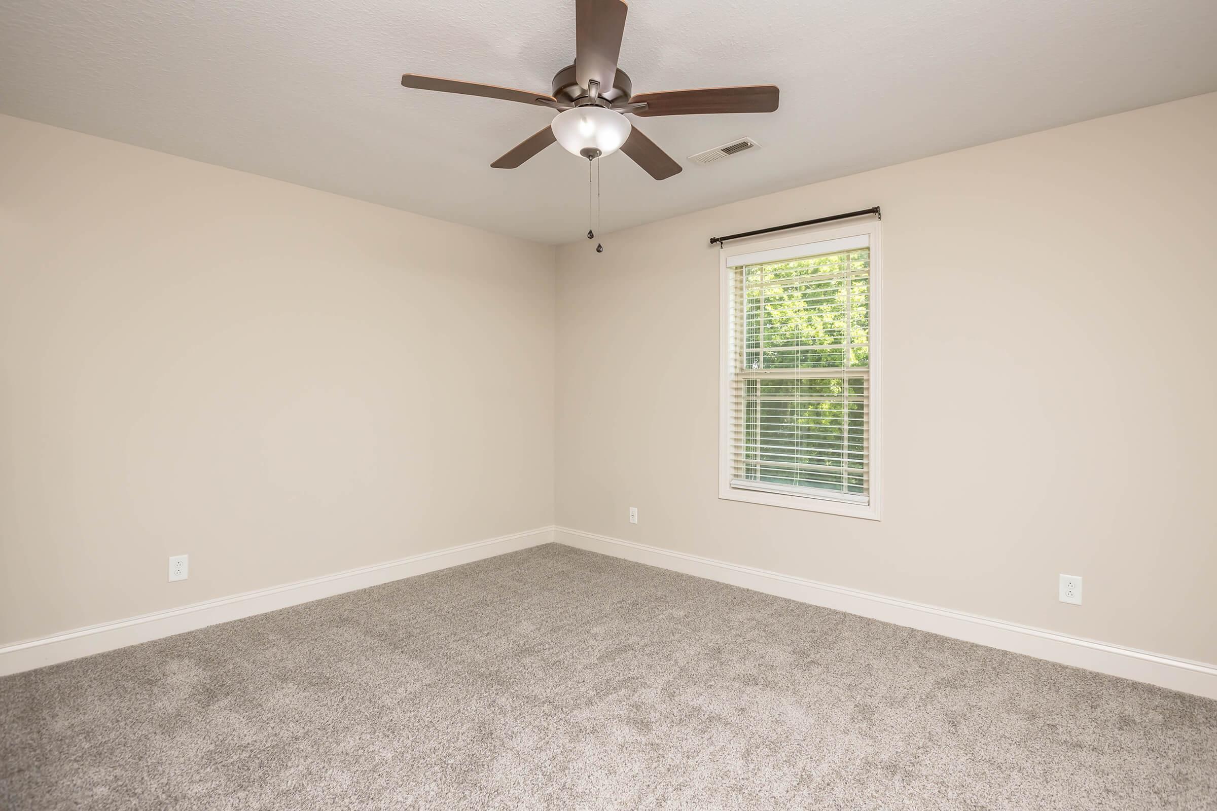 A neutral-colored empty bedroom featuring light beige walls, plush gray carpeting, a ceiling fan with wooden blades, and a window with white blinds allowing natural light. The room is spacious and uncluttered, ideal for personal decoration and setup.