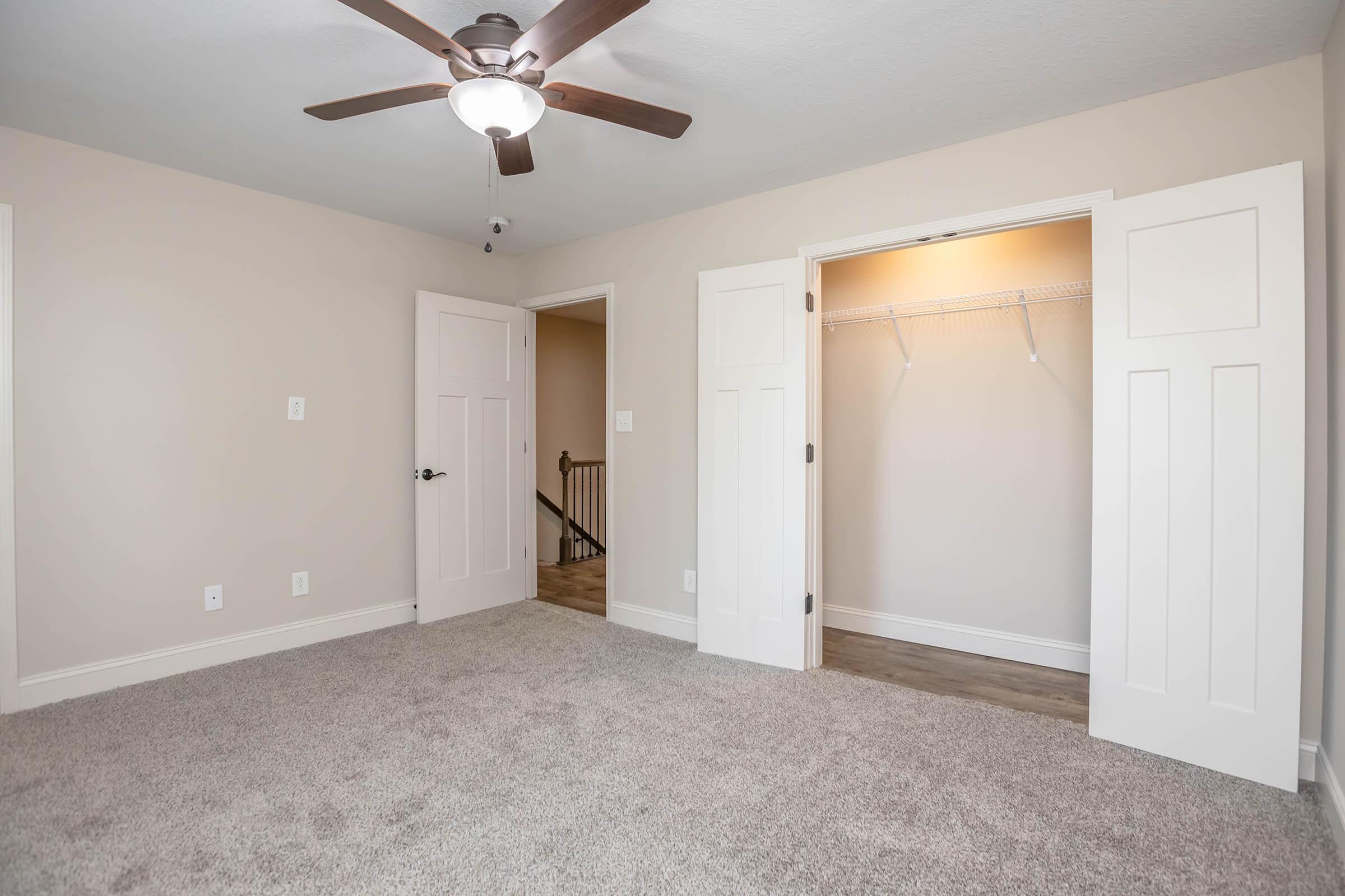A well-lit room featuring light beige walls and a ceiling fan. The floor is covered in carpet, and there are open double doors leading to a closet with a hanging rod. A staircase is visible in the hallway through the doorway. The overall atmosphere is spacious and inviting.
