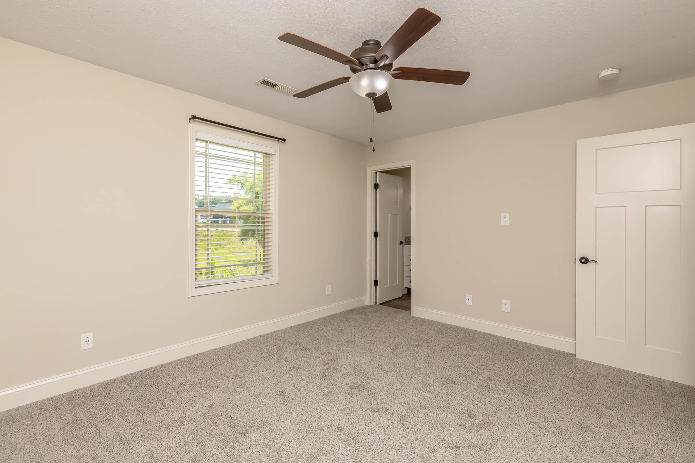 A spacious room with light beige walls and plush carpet. It features a ceiling fan, a window with white blinds allowing natural light, and a closed door leading to another room. The room is empty, creating a clean, minimalist aesthetic.