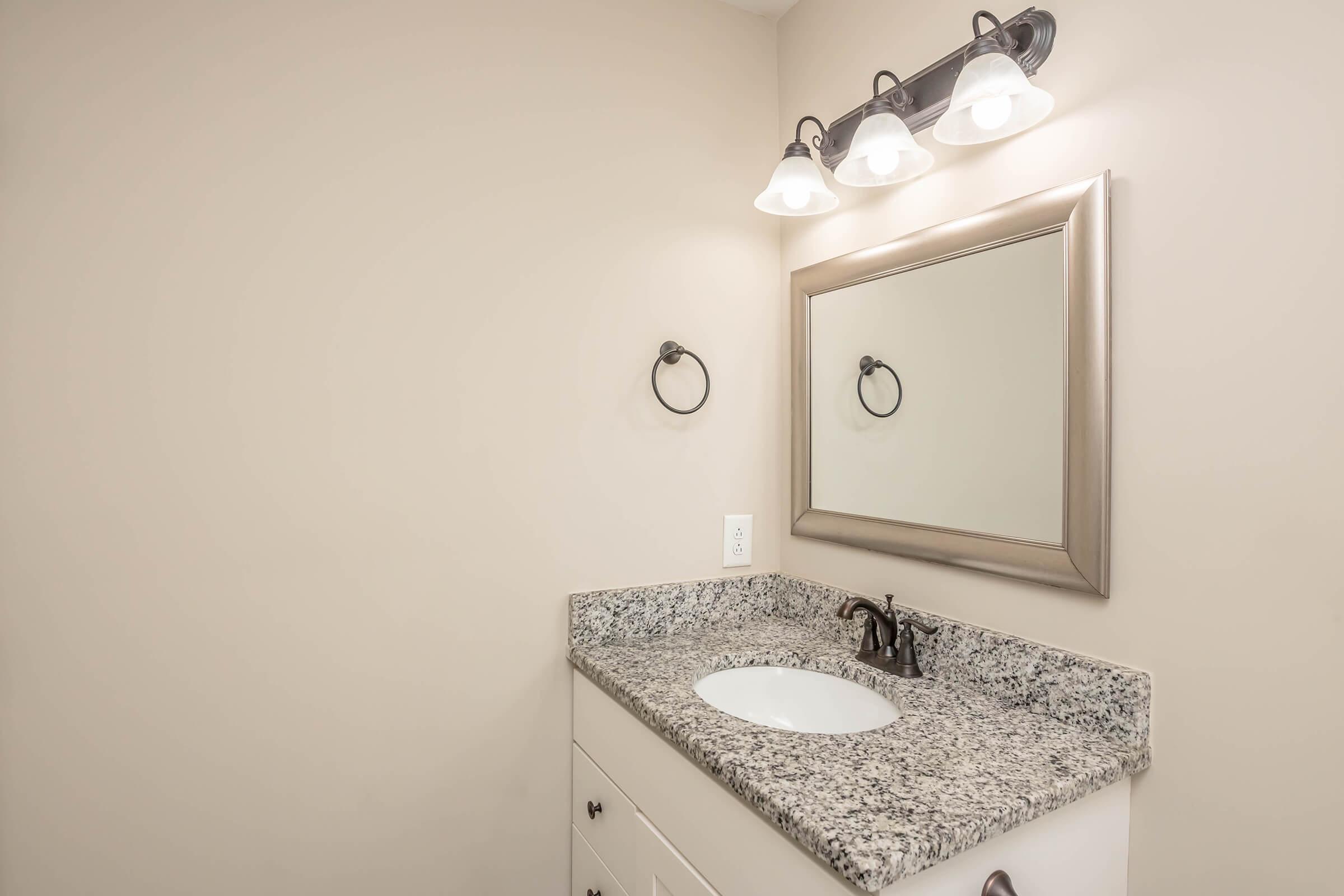 A modern bathroom featuring a light-colored wall, a granite countertop with an undermount sink, a decorative wall mirror framed in silver, and three light fixtures above the mirror. A towel ring is mounted next to the sink, creating a stylish and functional space.