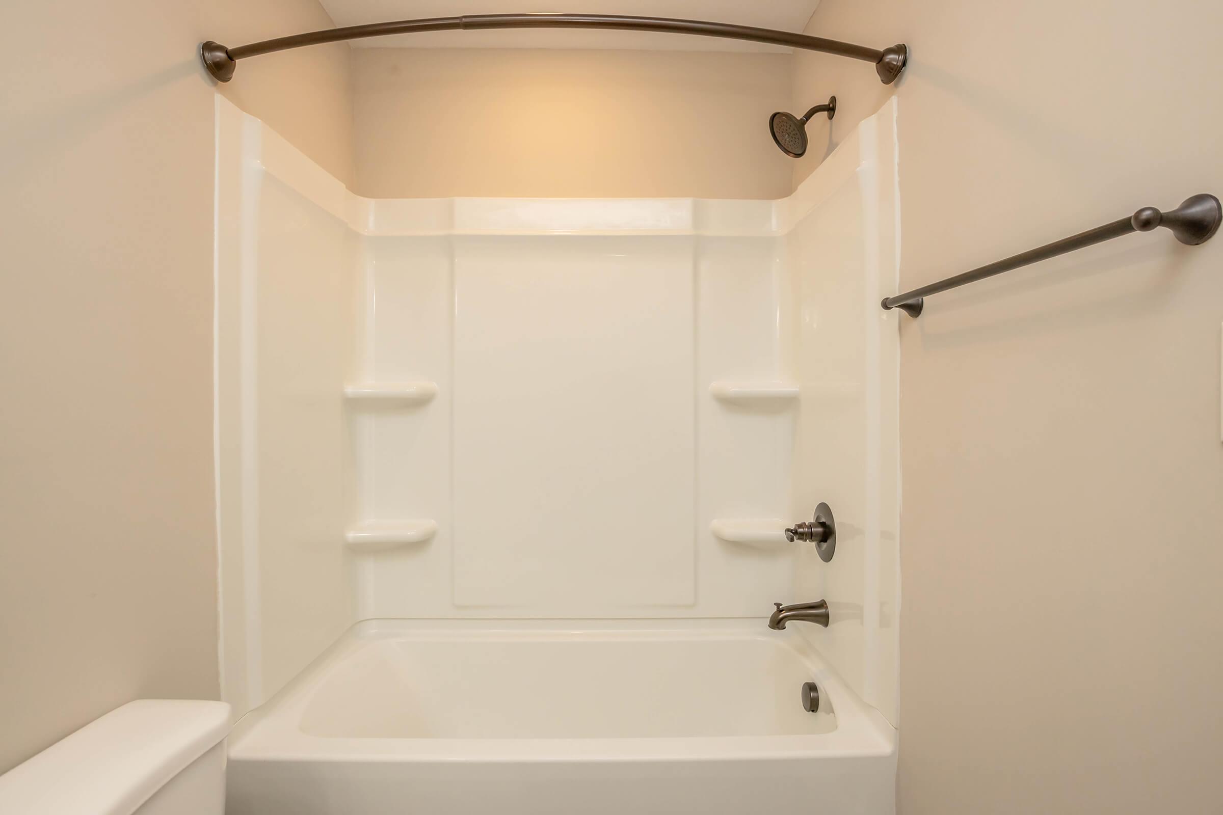 A clean, empty bathroom with a white acrylic bathtub and shower combination. The walls are smooth and beige, featuring a mounted showerhead and a towel bar. There are built-in shelves for toiletries. The space is well-lit with a soft overhead light.