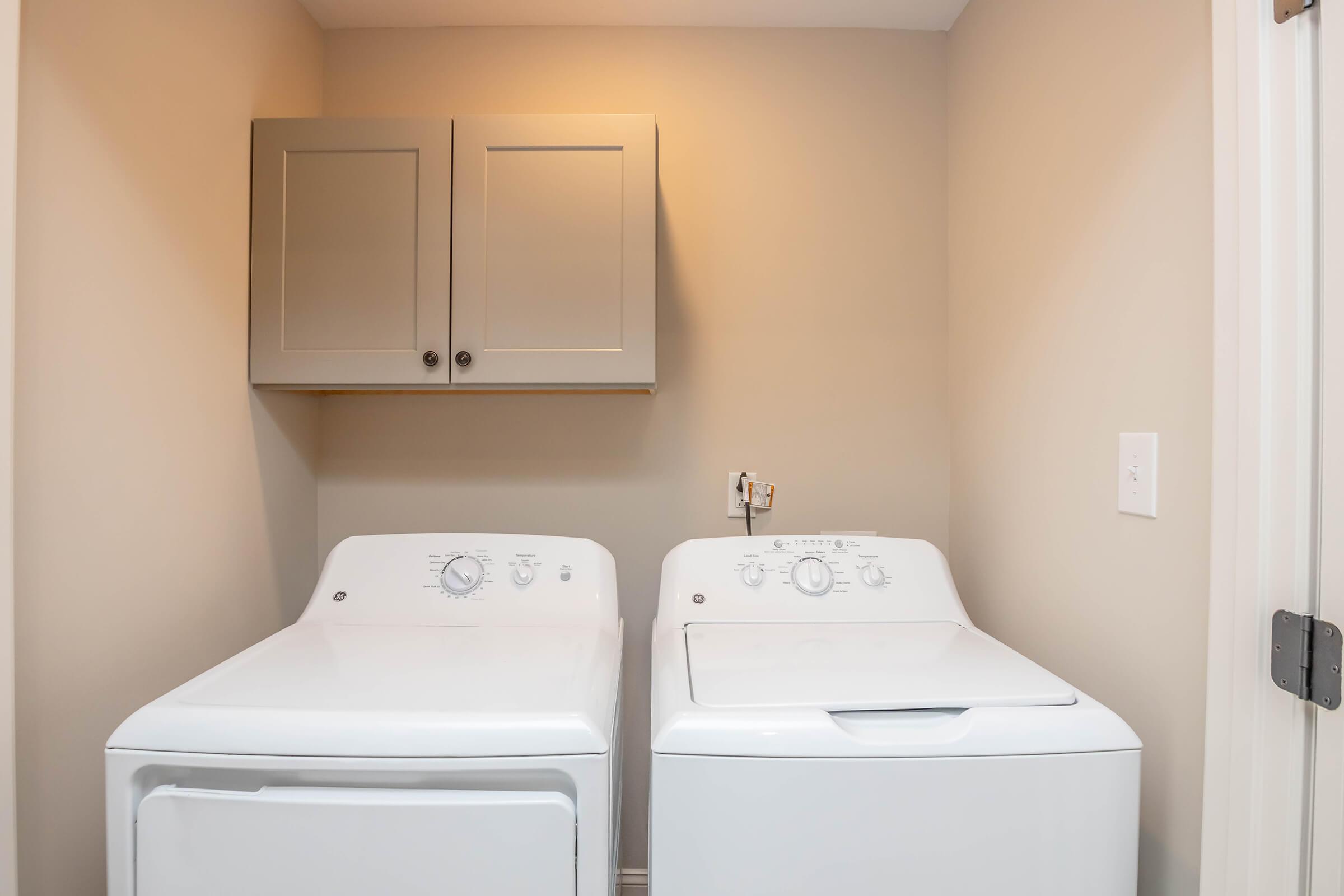 A laundry room featuring a paired washing machine and dryer in white, positioned side by side. Above them, there are two grey cabinets mounted on the wall. The room has neutral-colored walls and a door visible on the right side. The overall atmosphere is clean and organized.
