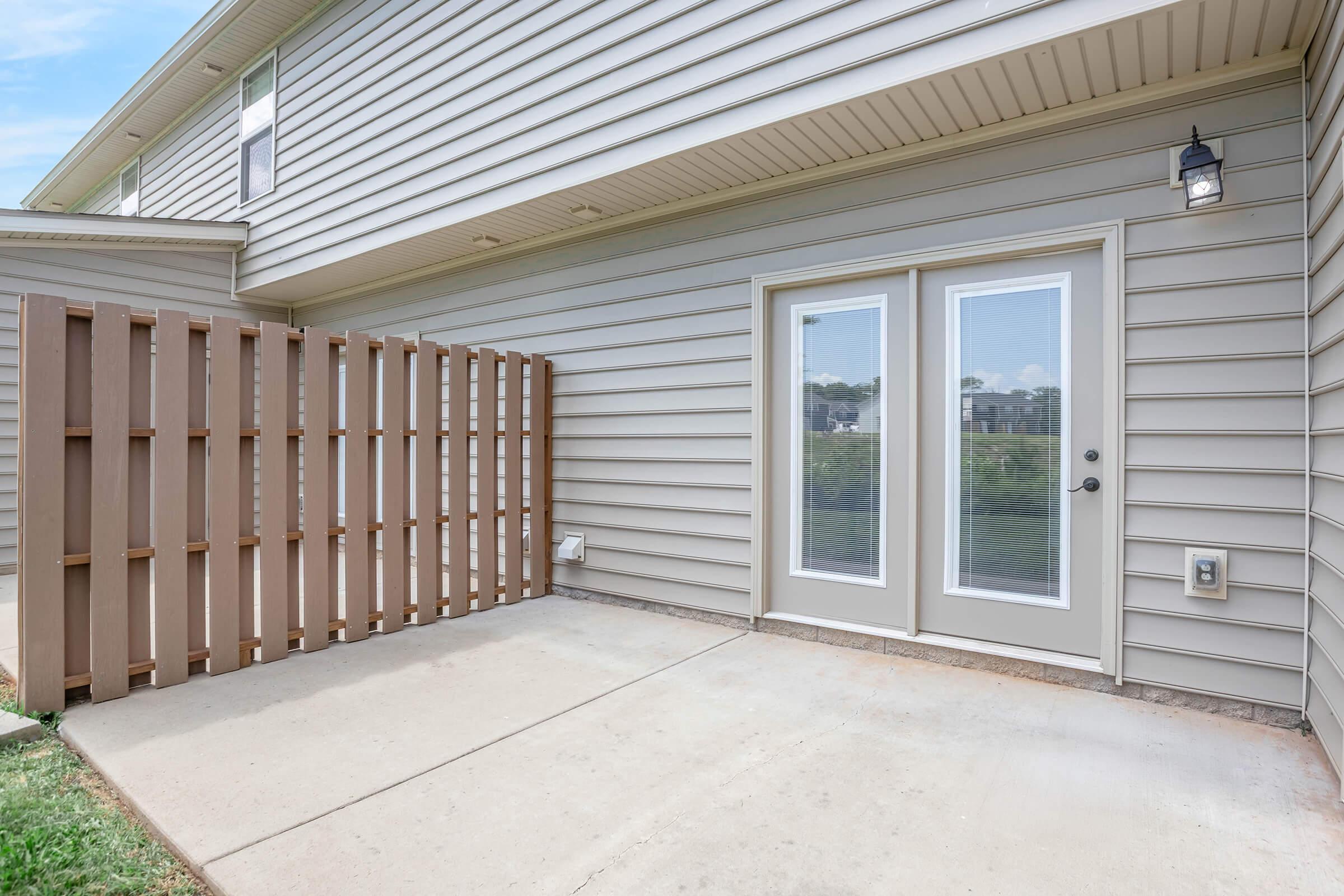 Patio area featuring a beige siding exterior, a pair of glass French doors with a dark door handle, and a wooden lattice fence. The ground is concrete, and there's a small light fixture next to the doors. Green grass and a view of surrounding buildings are visible in the background.