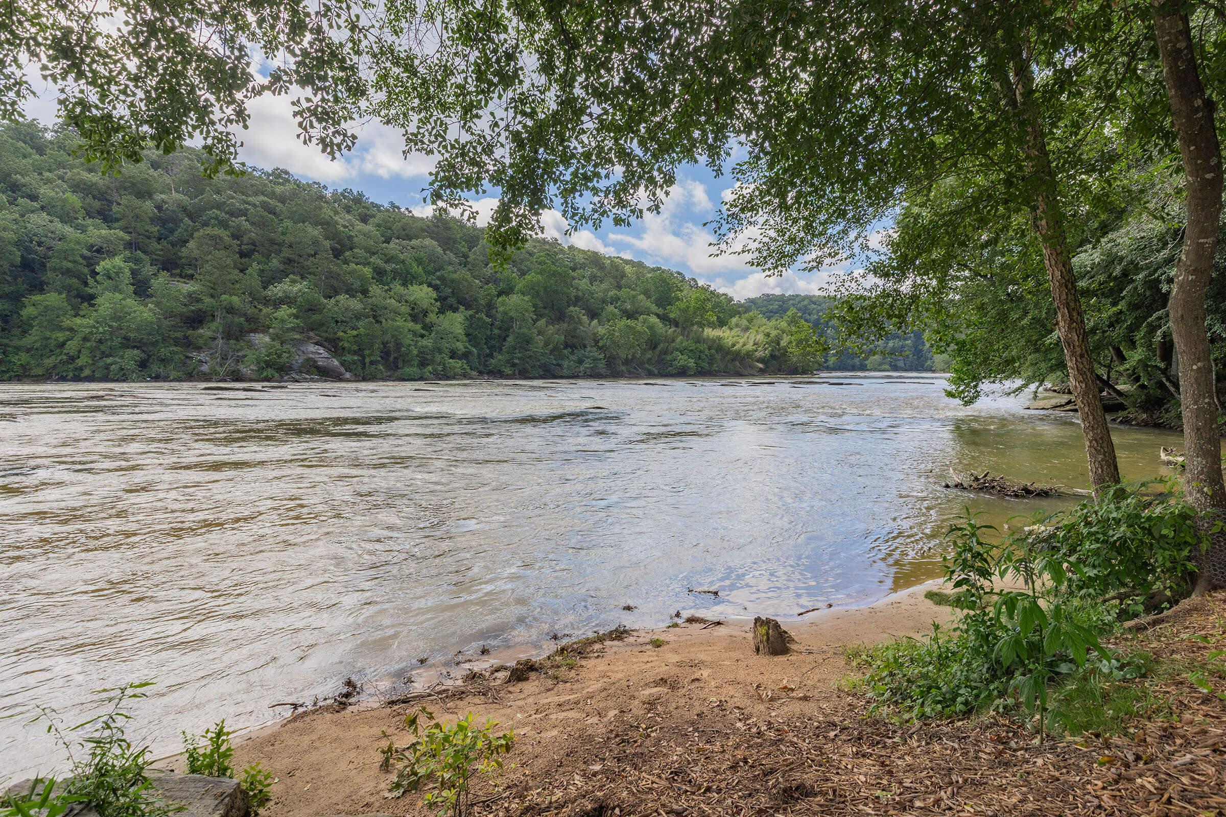 A scenic riverbank view featuring calm waters surrounded by lush greenery and trees. The landscape includes gentle hills in the background, providing a tranquil atmosphere with partly cloudy skies above. Perfect for outdoor activities or peaceful reflection by the water.