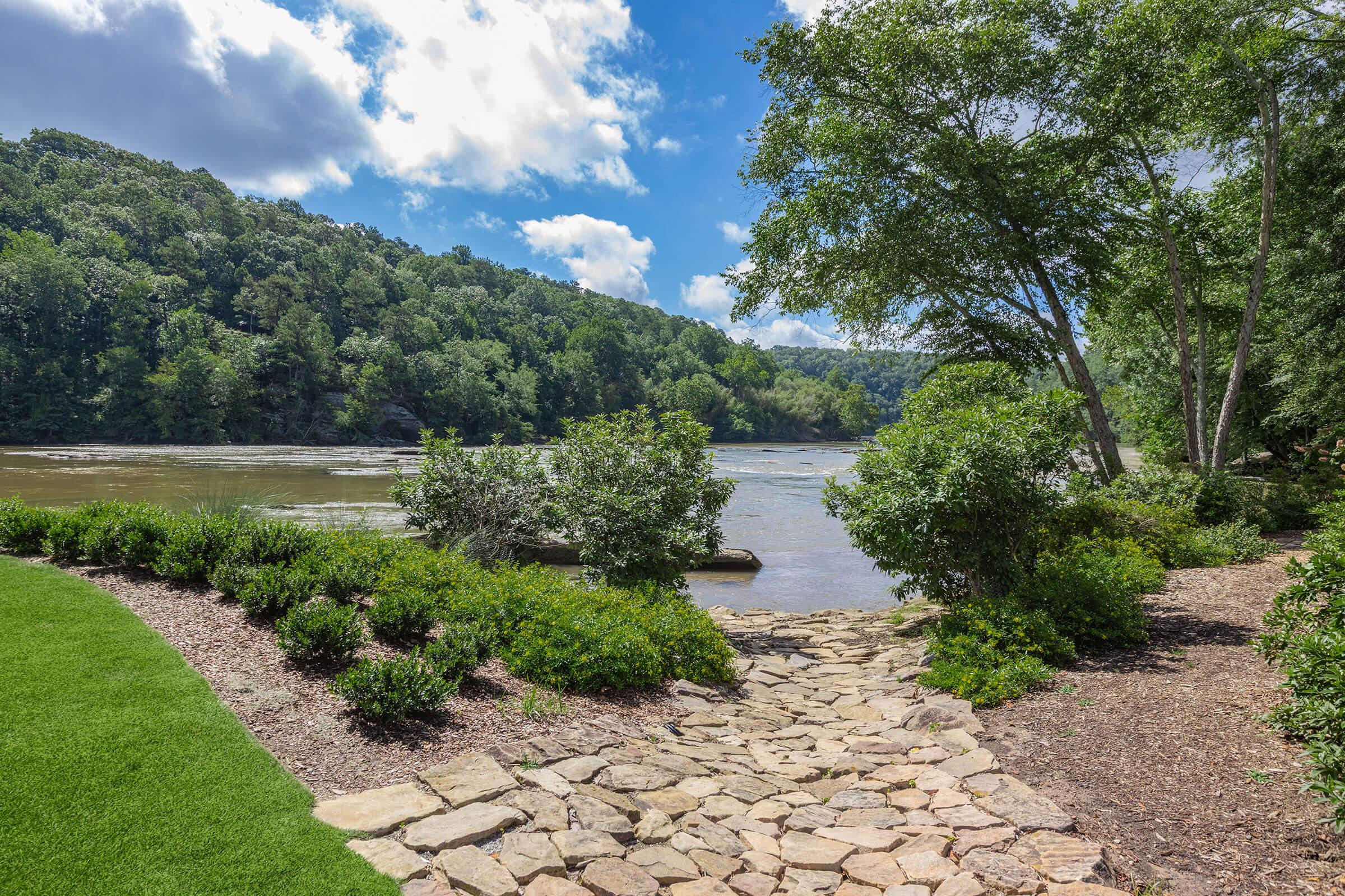A serene riverside scene featuring a stone pathway winding through lush greenery, leading to a calm river. The landscape is surrounded by trees and hills under a bright blue sky with fluffy clouds.