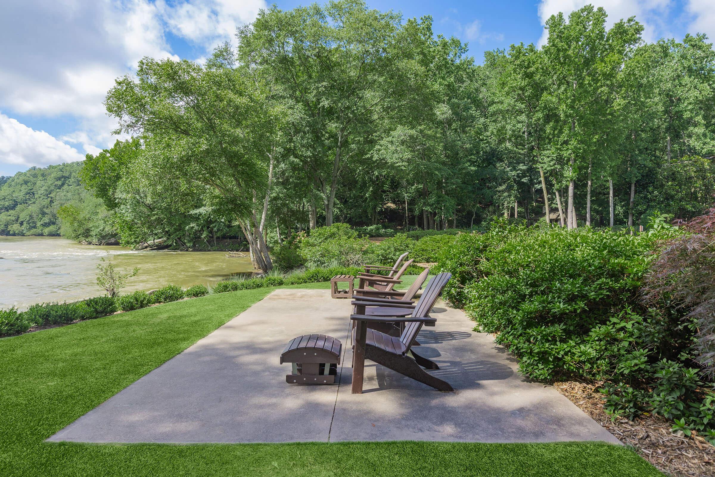 A serene riverside scene featuring multiple wooden Adirondack chairs arranged on a concrete patio, surrounded by lush greenery and trees. The calm river flows nearby, and the sky is partly cloudy, creating a peaceful outdoor setting perfect for relaxation.