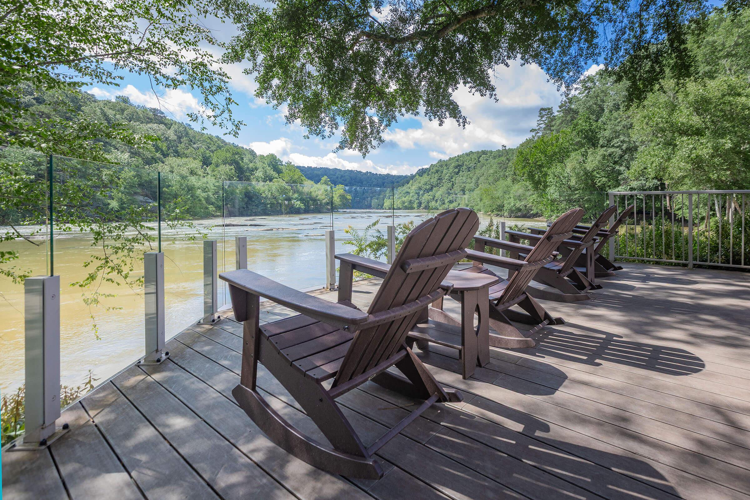 A serene riverside view featuring several wooden Adirondack chairs arranged on a deck. Lush green trees line the riverbank under a bright blue sky with fluffy white clouds, creating a peaceful outdoor setting perfect for relaxation.