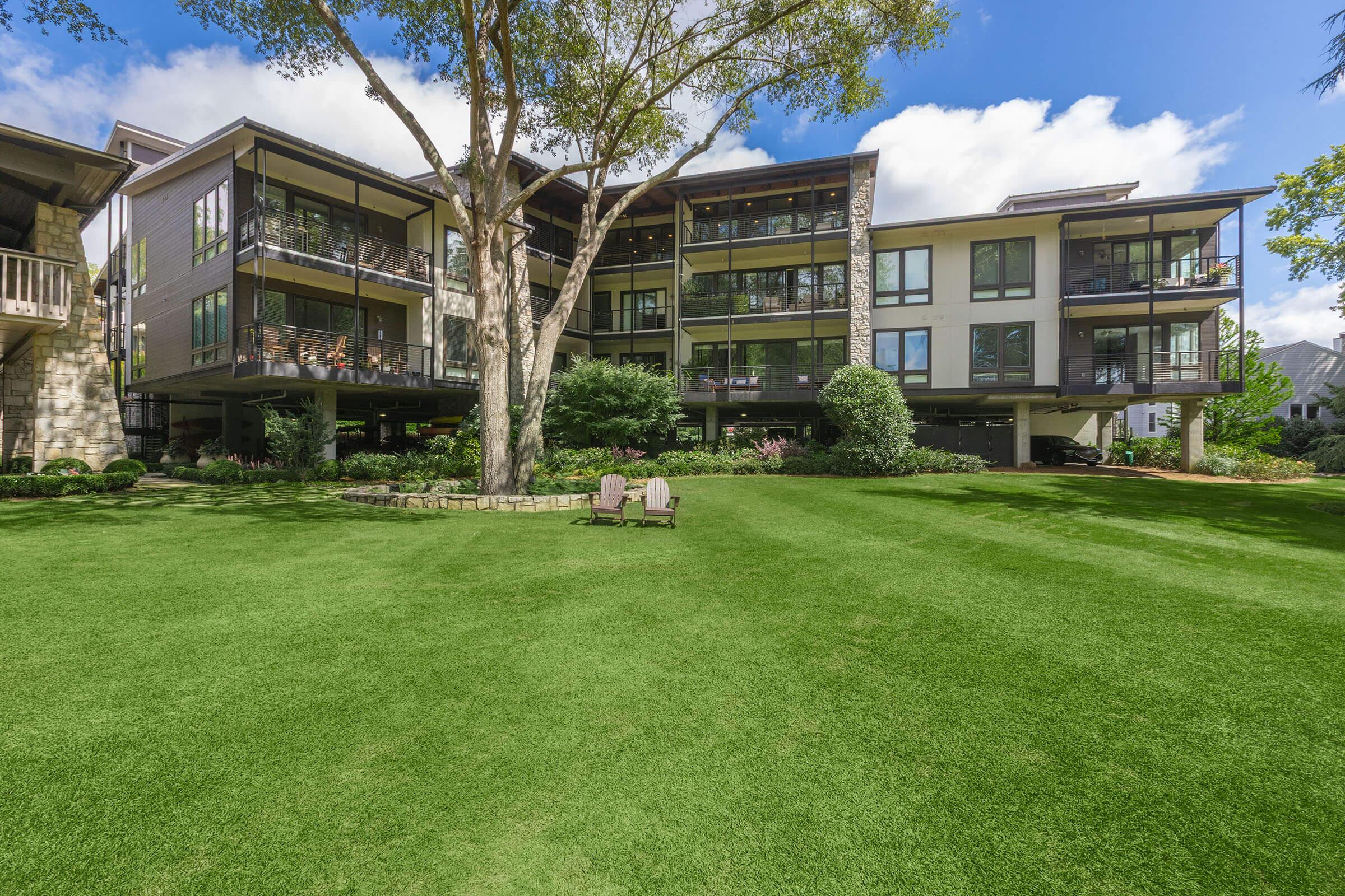 A modern multi-story building surrounded by lush green grass and trees. The building features large balconies and multiple windows, with outdoor seating areas on the lawn. Clear blue skies and fluffy clouds complete the serene, inviting landscape.