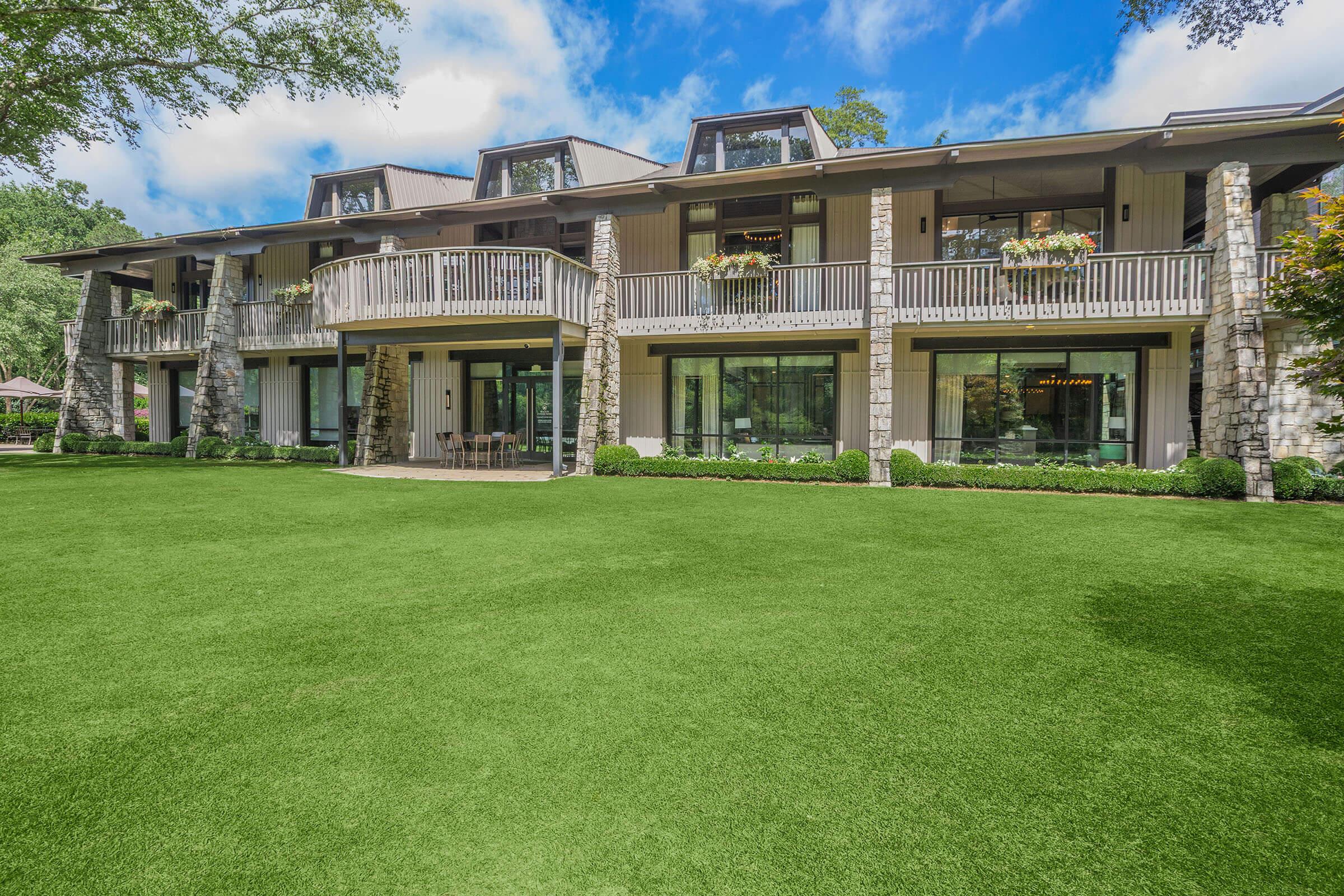 Exterior view of a modern house featuring large windows and multiple balconies. The building is surrounded by well-maintained landscaping, with a lush green lawn in the foreground. The sky is partly cloudy, adding to the serene atmosphere of the setting.