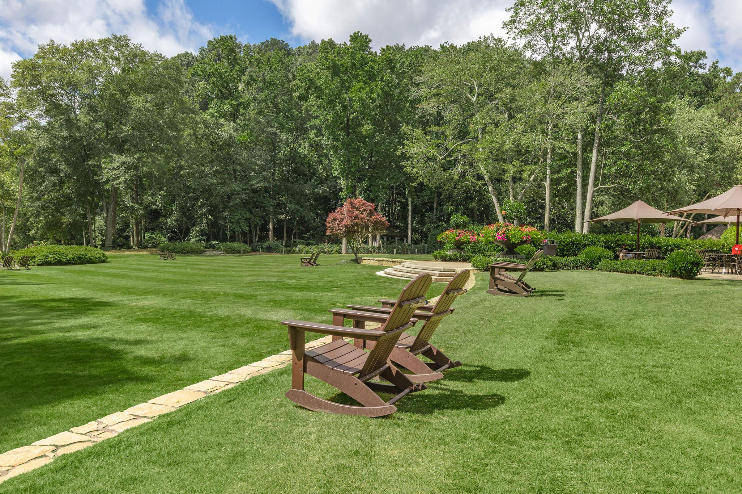 A serene outdoor space featuring well-maintained green grass, elegant wooden Adirondack chairs arranged in a pleasant setting. Lush trees and colorful plants surround the area, with stone pathways leading towards a beautiful pavilion in the background, creating a peaceful retreat in nature.