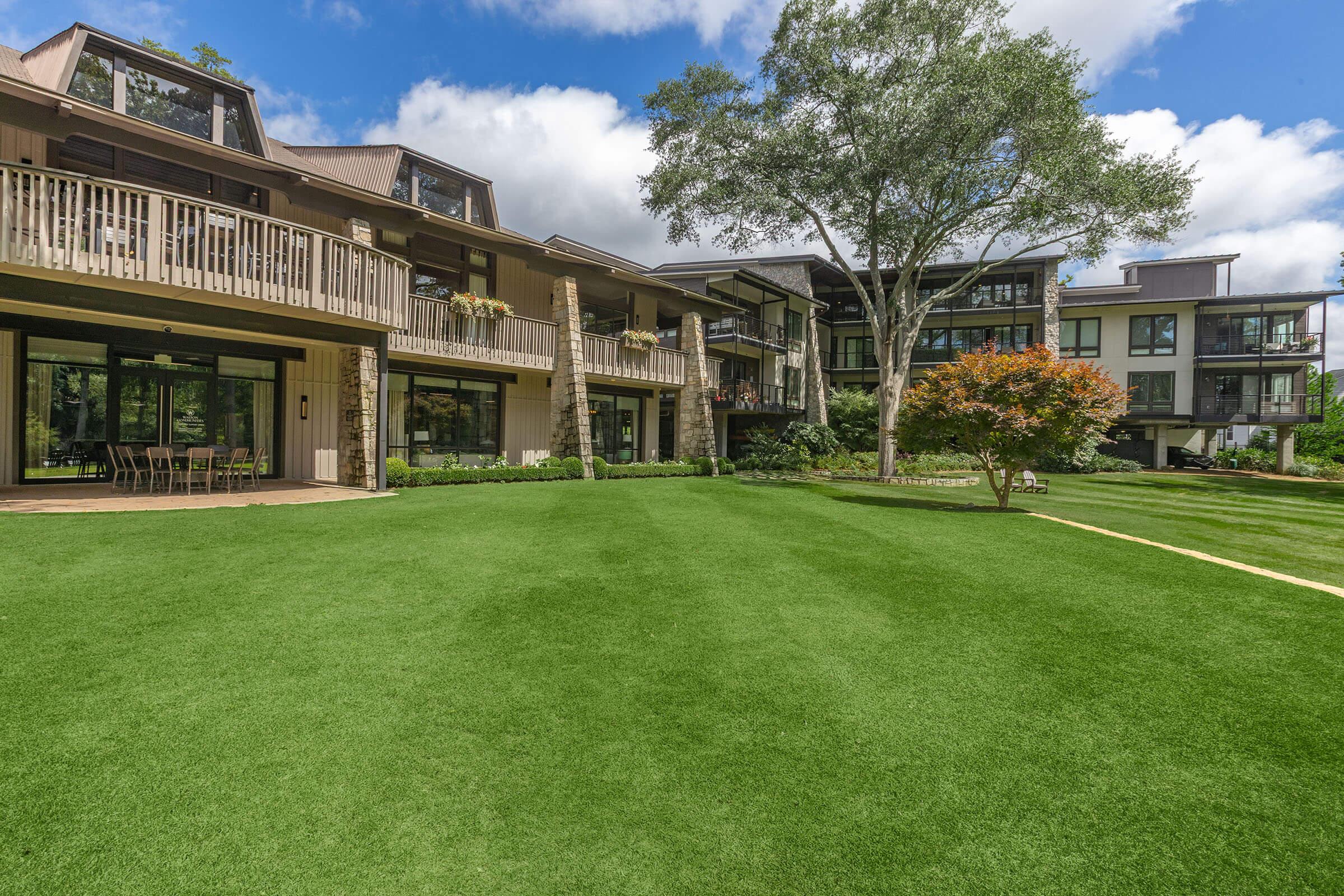 A modern residential building with multiple levels, featuring large windows and balconies, surrounded by a well-maintained green lawn. A tree and a small decorative tree add to the landscape, under a bright blue sky with scattered clouds.