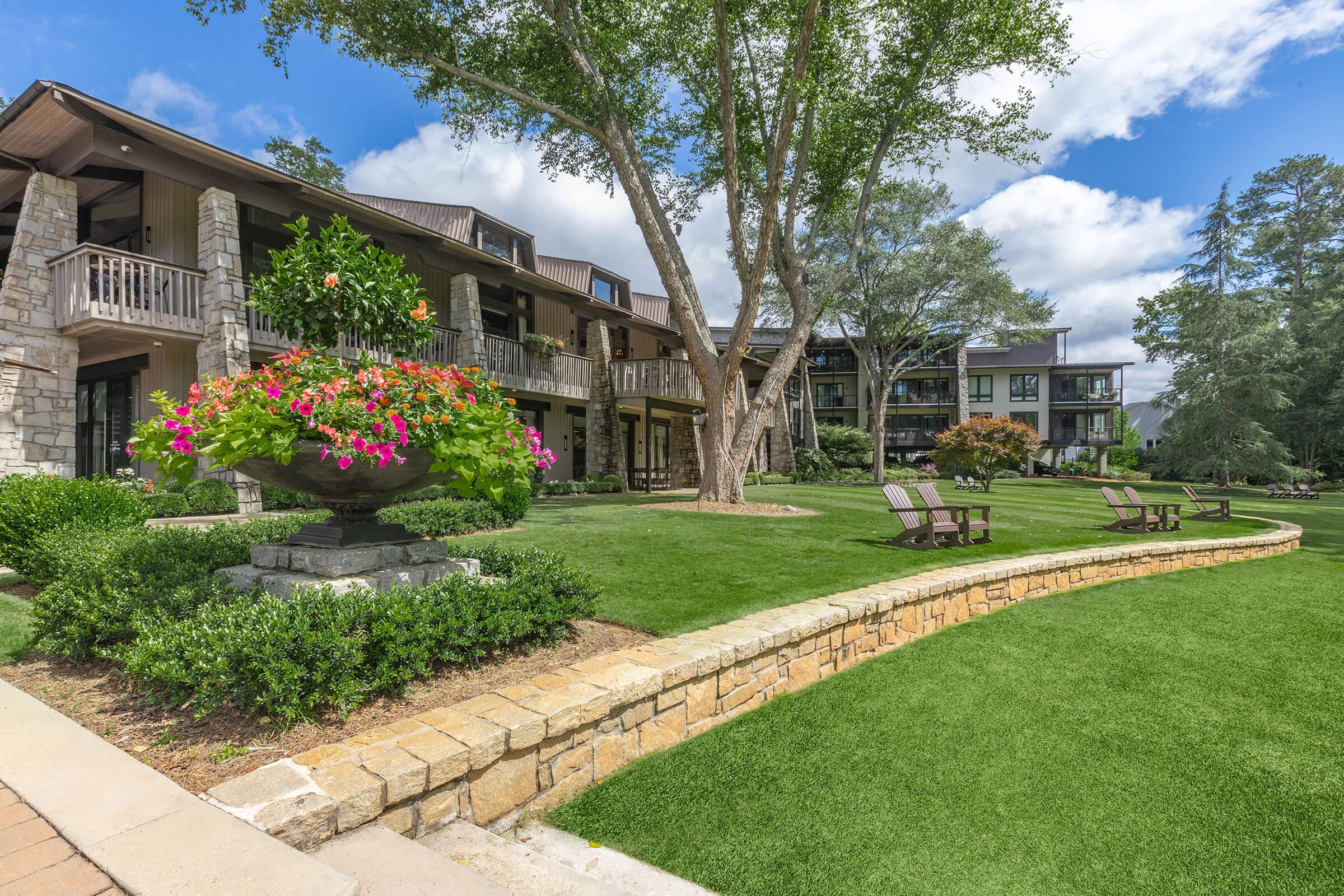 A beautifully landscaped area featuring a well-maintained lawn, flower-filled planters, and seating arrangements. Two wooden lounge chairs sit on the grass. A multi-story building with a stone exterior and balconies is visible in the background, under a blue sky with scattered clouds.