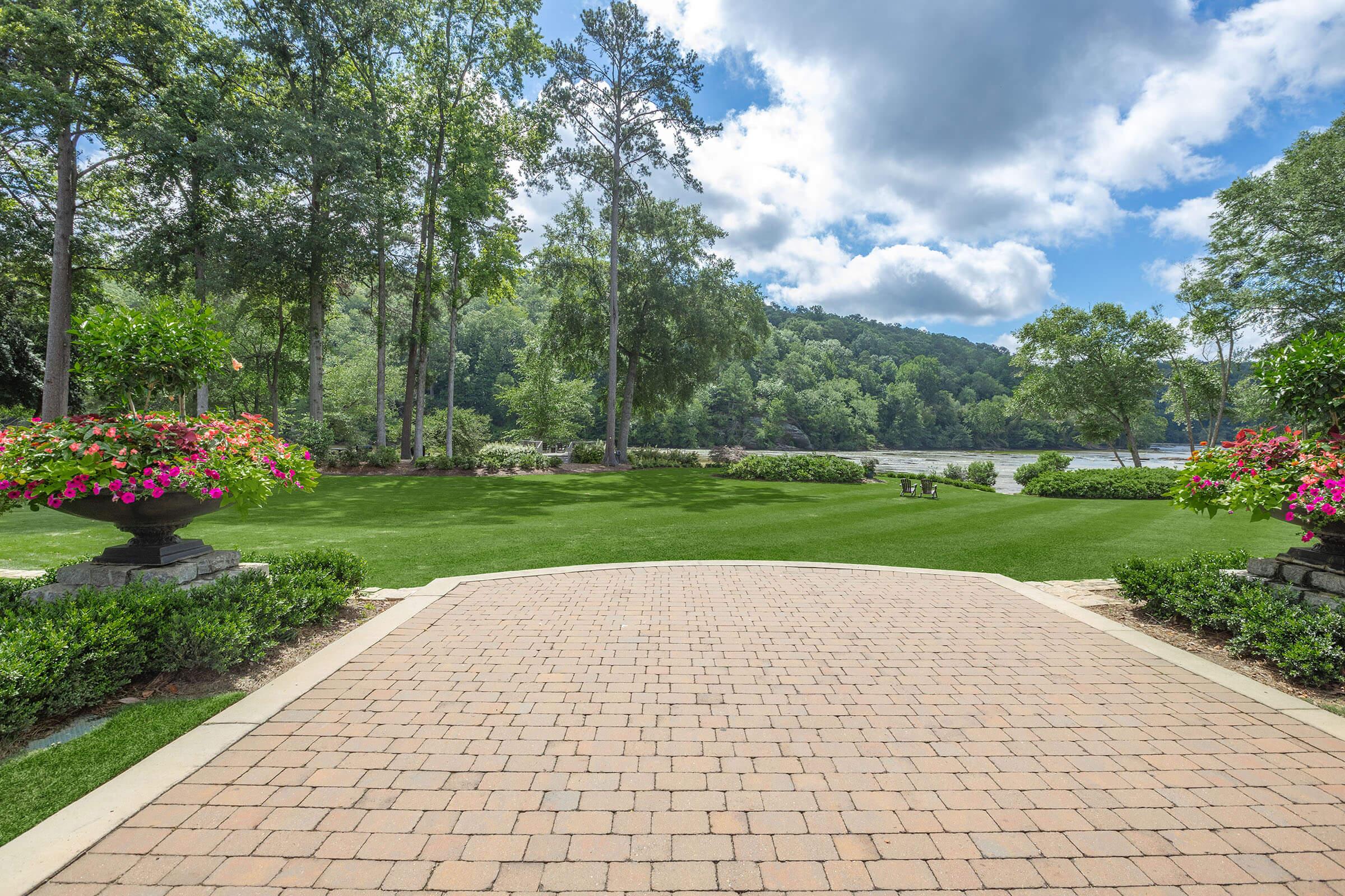 A picturesque landscape featuring a paved pathway leading to a lush green lawn bordered by flowering plants. Tall trees provide shade, and a serene lake is visible in the background under a partly cloudy sky. The scene evokes tranquility and natural beauty.