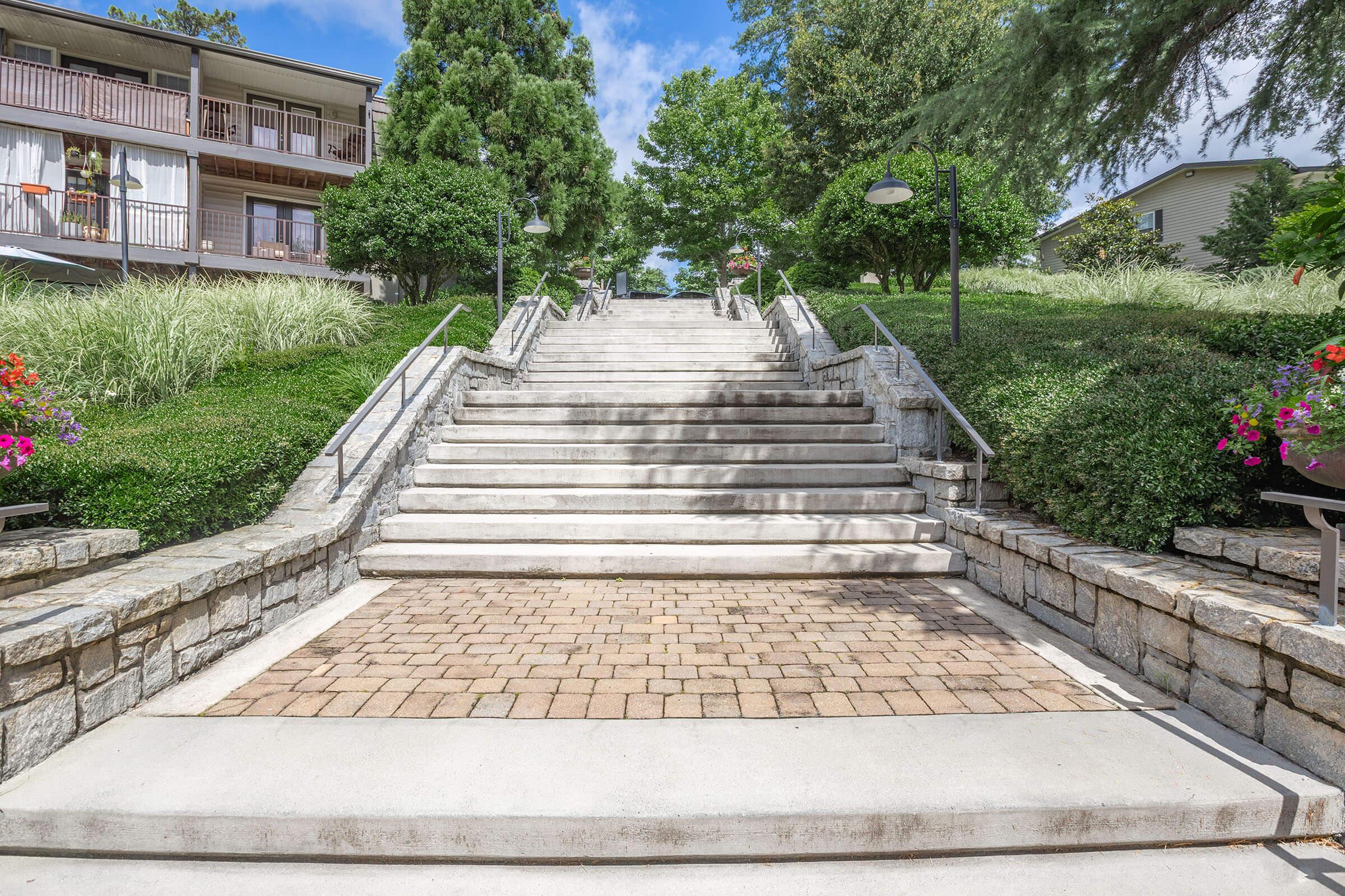 A set of wide stone stairs flanked by lush greenery and flowering plants, leading upward in a landscaped area. The stairs are made of concrete, and there are handrails on both sides. In the background, there are buildings with balconies surrounded by trees and clouds in the blue sky.