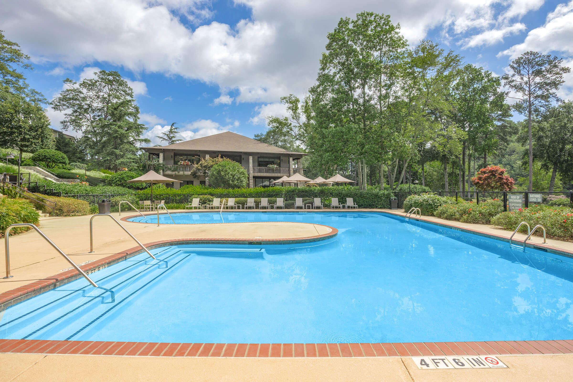 A bright, inviting swimming pool surrounded by lush greenery and lounge chairs. The pool has clear blue water and features a gradual entry. In the background, a spacious building with a patio and shaded areas is visible under a partly cloudy sky.
