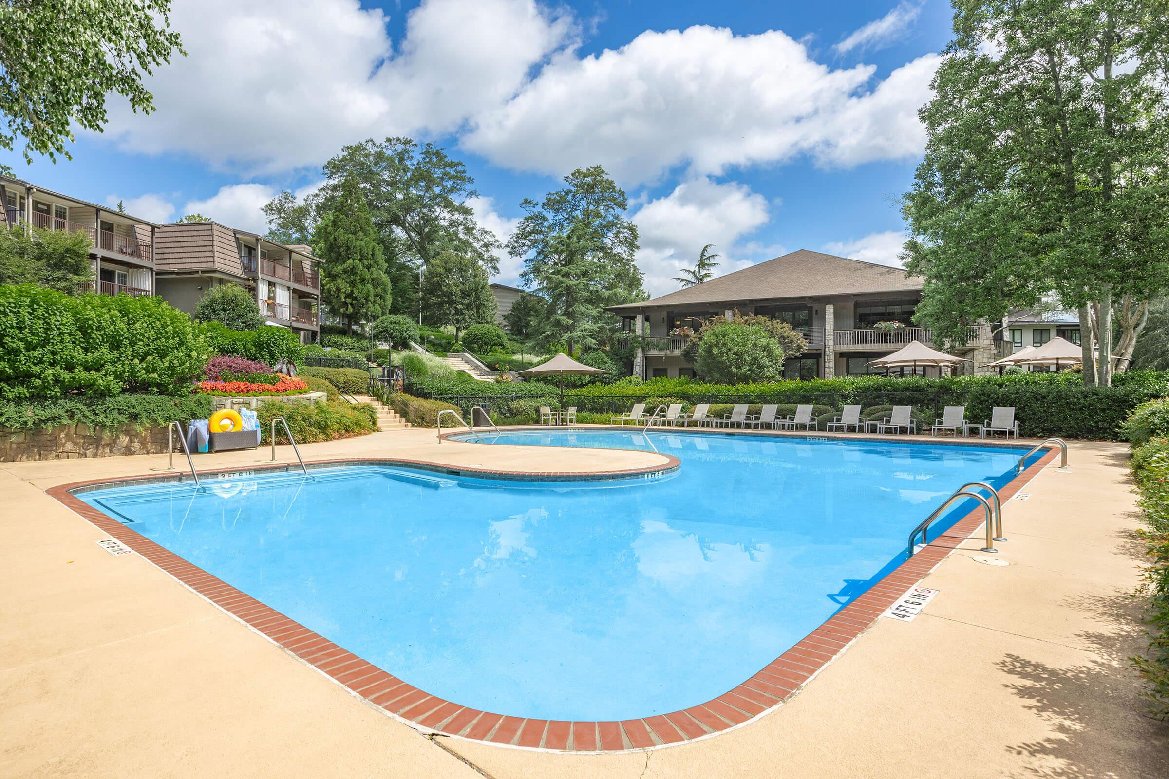 A clear blue swimming pool surrounded by lush greenery and neatly arranged lounge chairs. The area is sunny with fluffy clouds overhead, and there are umbrellas and landscaped gardens in the background, creating a serene and inviting atmosphere.