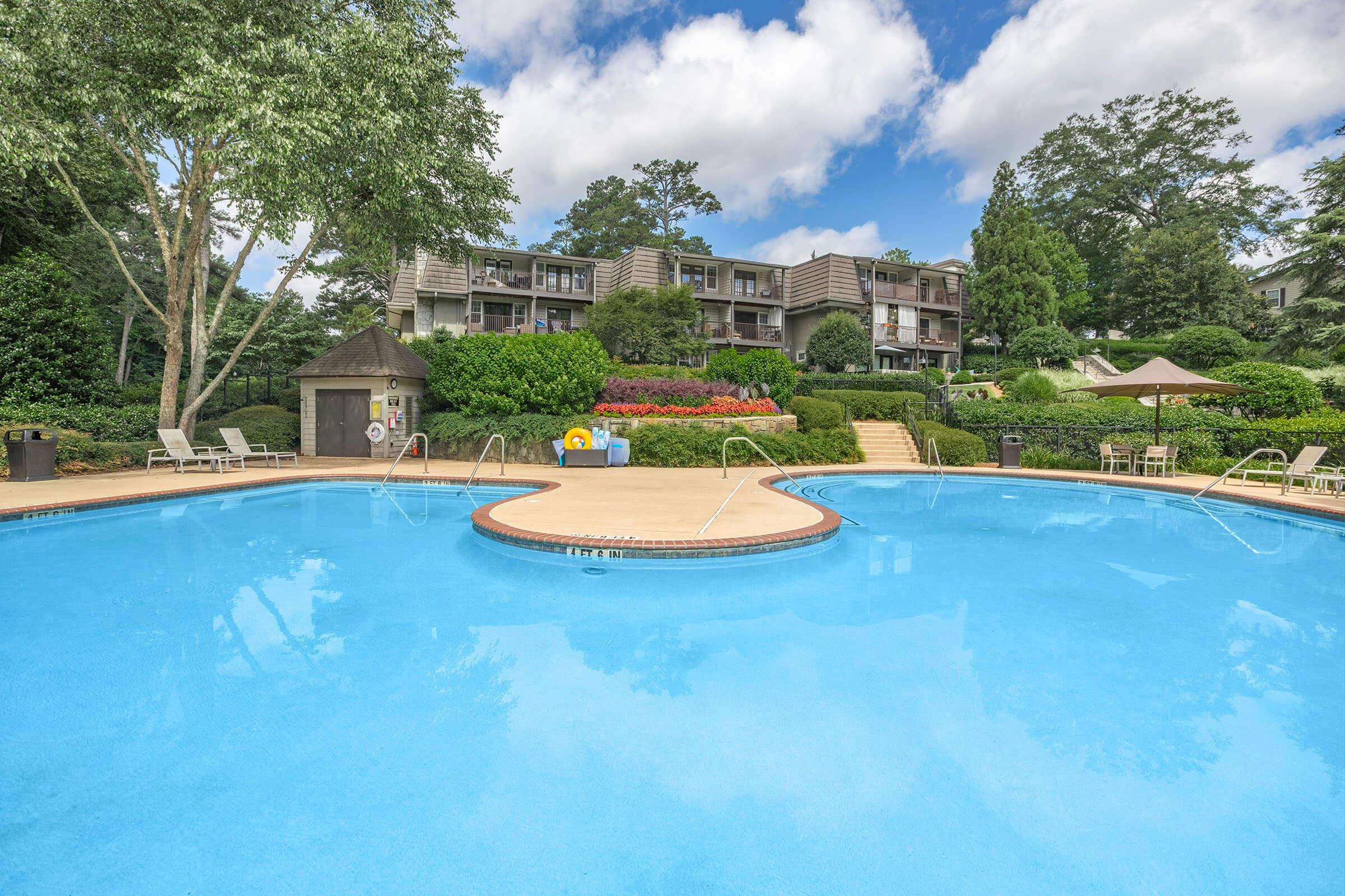 A serene outdoor pool surrounded by lush greenery and colorful blooming plants. In the background, there are multi-story buildings with balconies. The sky is partly cloudy, creating a peaceful atmosphere. Sun loungers and umbrellas are placed around the pool area, enhancing the relaxation scenery.