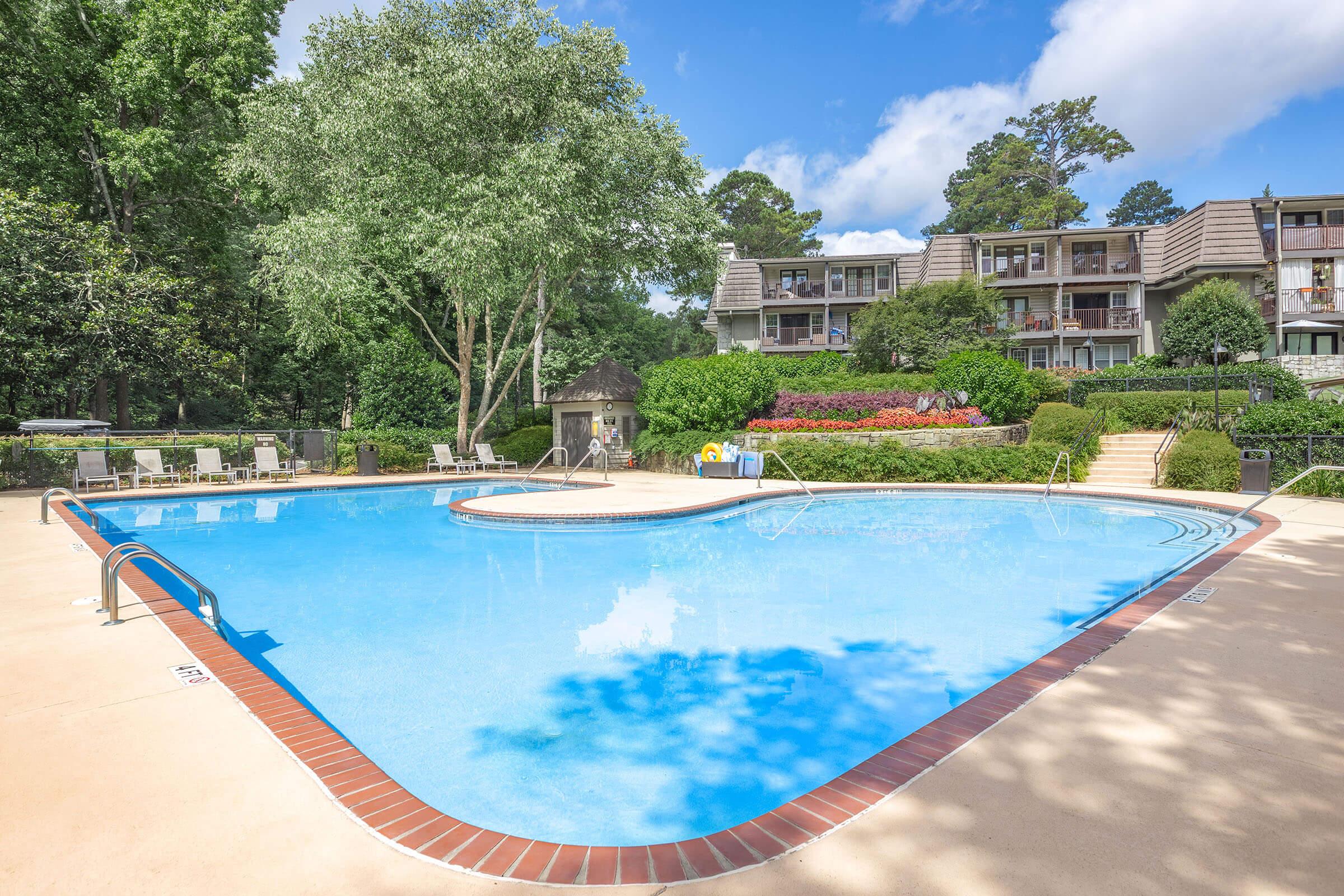 A clear, blue swimming pool surrounded by lush greenery and well-maintained landscaping. In the background are multi-story apartment buildings with balconies. There are lounge chairs near the pool, and vibrant flowers add color to the scene under a partly cloudy sky.