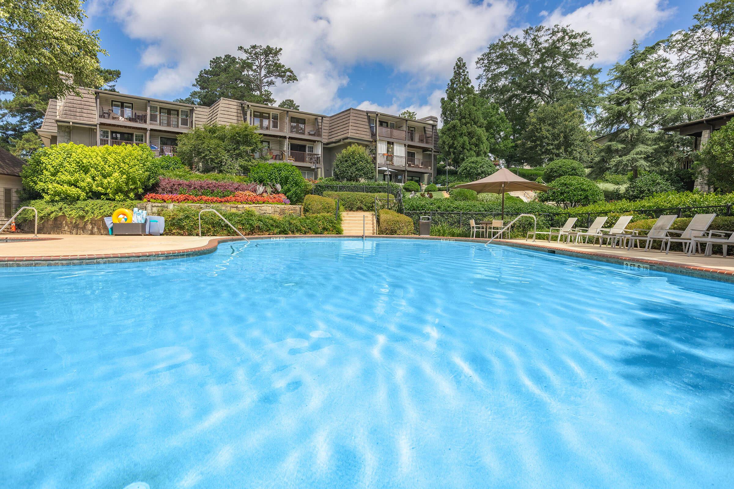 A clear blue swimming pool reflecting the sky, surrounded by lush greenery and colorful flower beds. In the background, there are several multi-story buildings with balconies. Poolside chairs are neatly arranged along the edge, and a sunshade structure is visible nearby, creating a serene outdoor atmosphere.