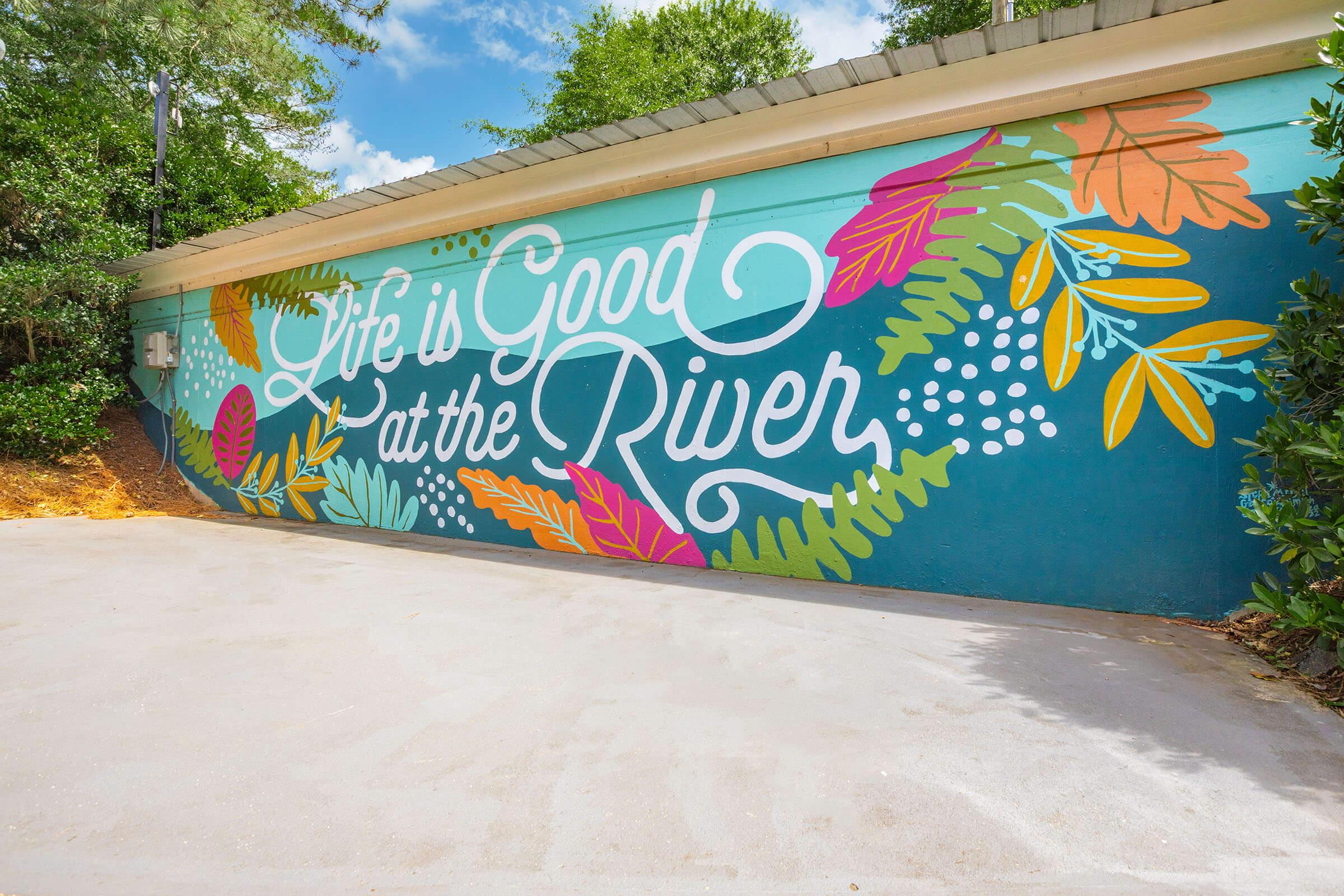 Colorful mural on a wall featuring the phrase "Life is Good at the River," surrounded by vibrant leaves and floral designs against a bright blue background. The setting is outdoors with trees and a clear sky visible.