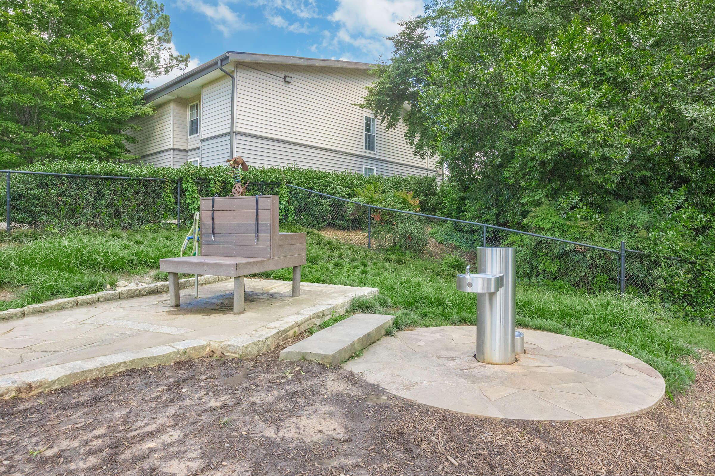 A small outdoor seating area features a wooden bench on a stone base, adjacent to a modern water fountain. The area is surrounded by grassy space and trees, with a residential building visible in the background. A fence encloses the space, creating a peaceful, park-like setting.