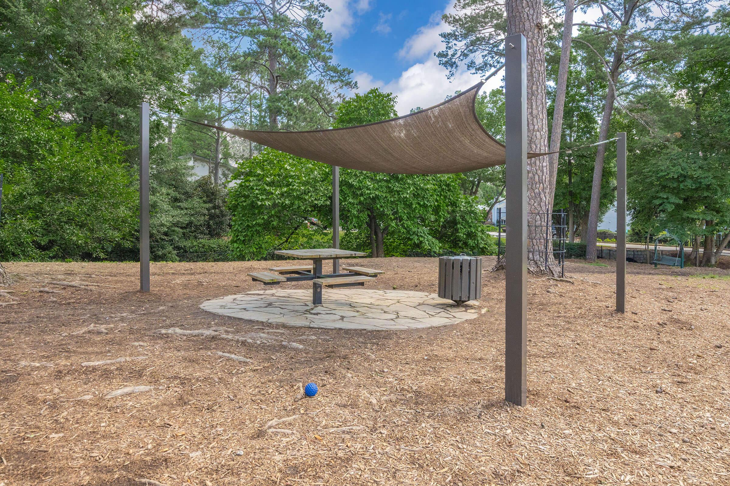 A shaded picnic area with a stone circular seating arrangement and a wooden table, surrounded by trees and green landscaping. The ground is covered with wood chips, and there is a small blue ball nearby. Bright skies are visible above, indicating a pleasant day.