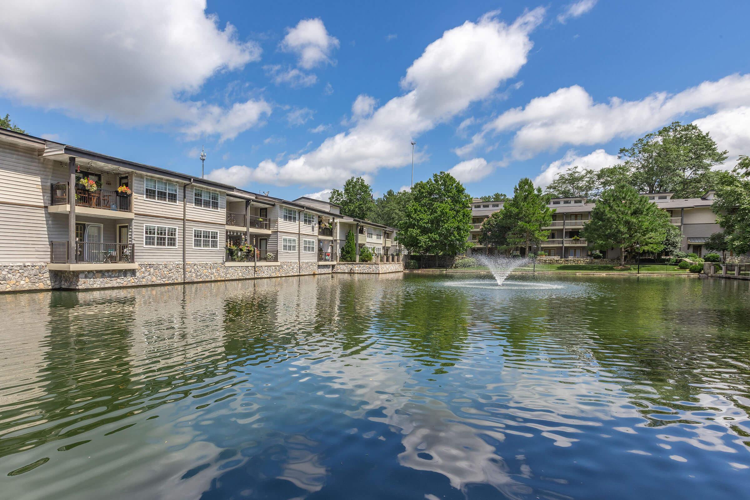 A serene view of a residential complex by a pond, featuring two buildings with balconies adorned with plants, surrounded by lush greenery. A fountain is centered in the pond, reflecting the blue sky and fluffy clouds above. The scene captures a peaceful, inviting atmosphere.
