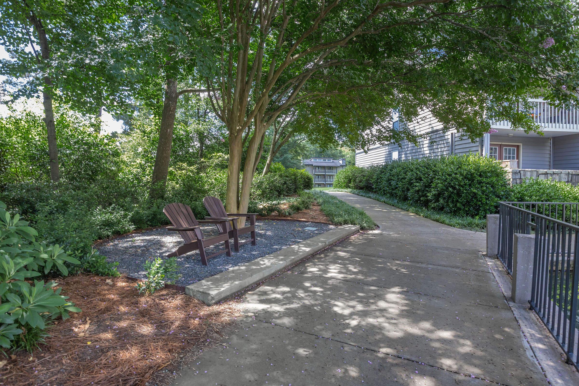 A serene outdoor pathway lined with greenery and bushes, featuring two wooden adirondack chairs under a large tree. The path is made of concrete and meanders next to a garden area, providing a peaceful spot to relax in nature. Sunlight filters through the leaves, casting dappled shadows on the ground.