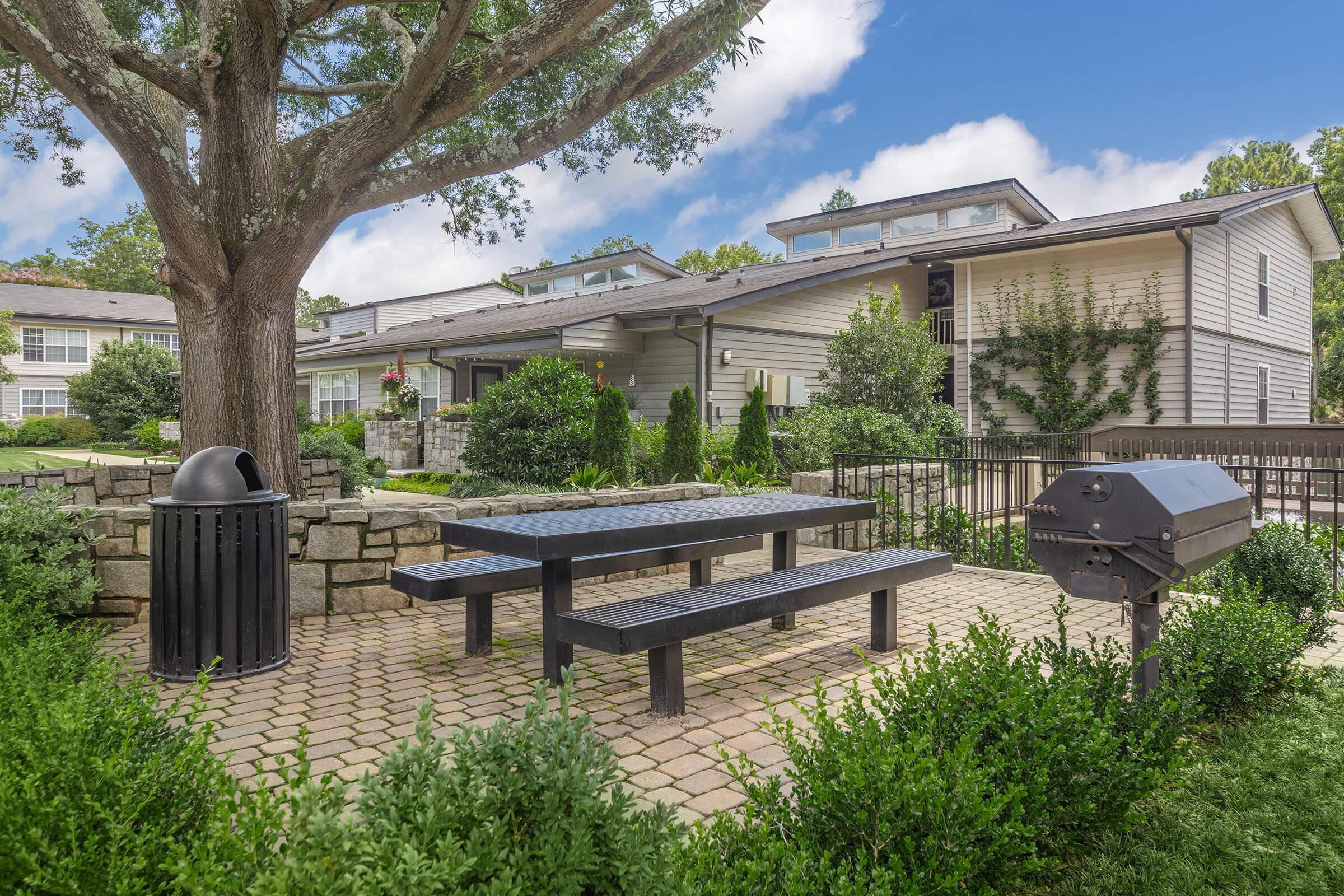 An outdoor picnic area with a black picnic table and grill surrounded by neatly trimmed bushes and trees. The setting features a stone walkway and provides a view of a modern building in the background. The space appears inviting and well-maintained, suitable for gatherings.