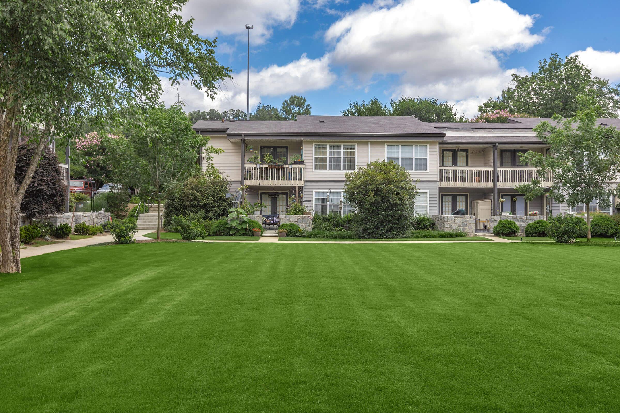 A view of a well-maintained residential complex surrounded by lush green lawns and trees. The buildings feature balconies, and there is a clear blue sky with clouds in the background, creating a serene and inviting atmosphere. Ideal for a peaceful living environment.